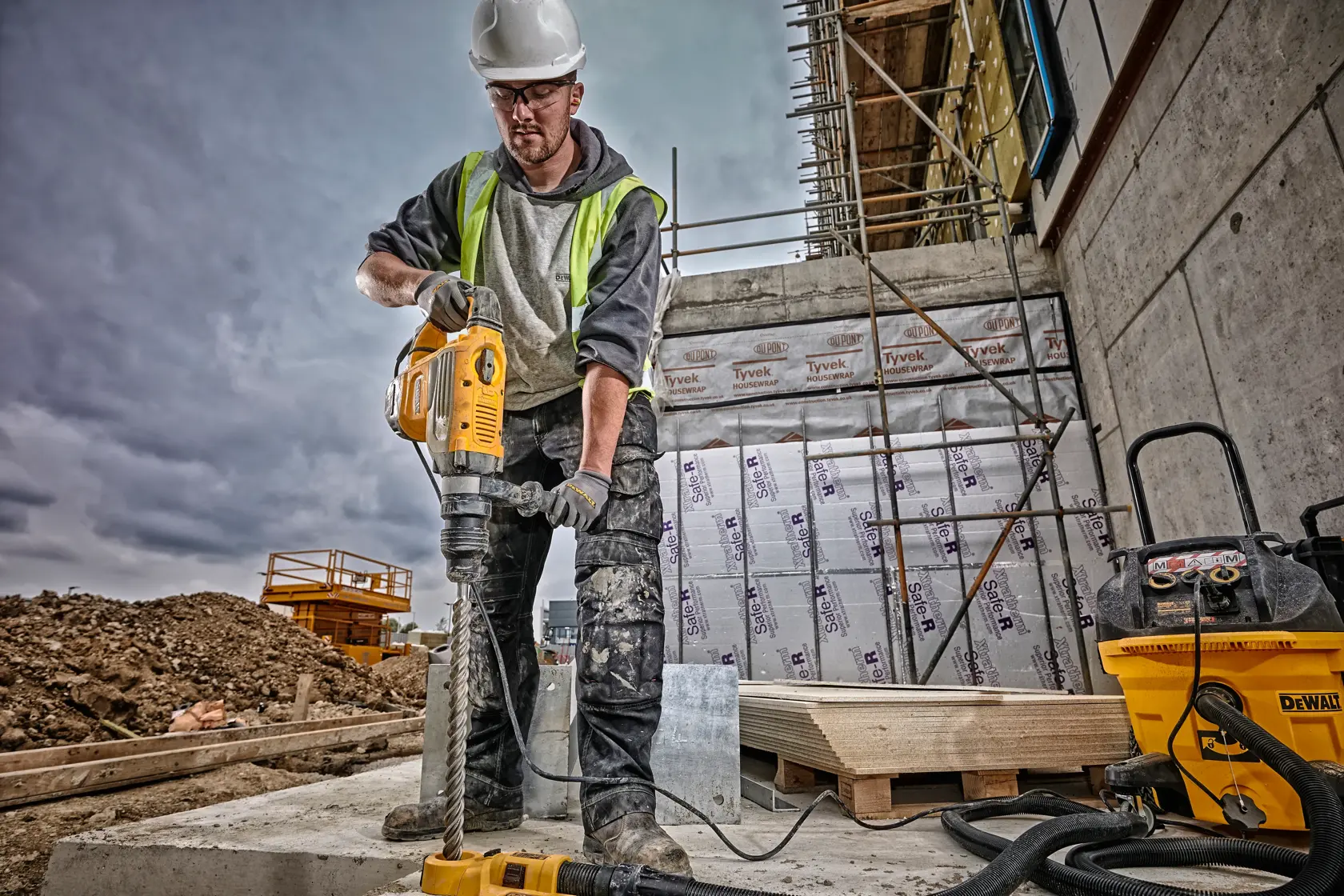 Un trabajador de la construcción con casco de seguridad y chaleco reflectante utiliza un martillo rotativo DEWALT (SKU: D25733K) en una obra. El rostro del trabajador está difuminado, y en el fondo hay equipos de construcción, andamios y materiales de aislamiento.