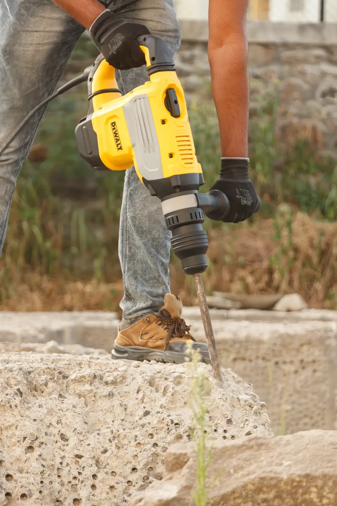Una persona usa un martillo rotatorio amarillo DEWALT (modelo D25733K) para romper concreto al aire libre. La persona lleva guantes, jeans y botas de trabajo.