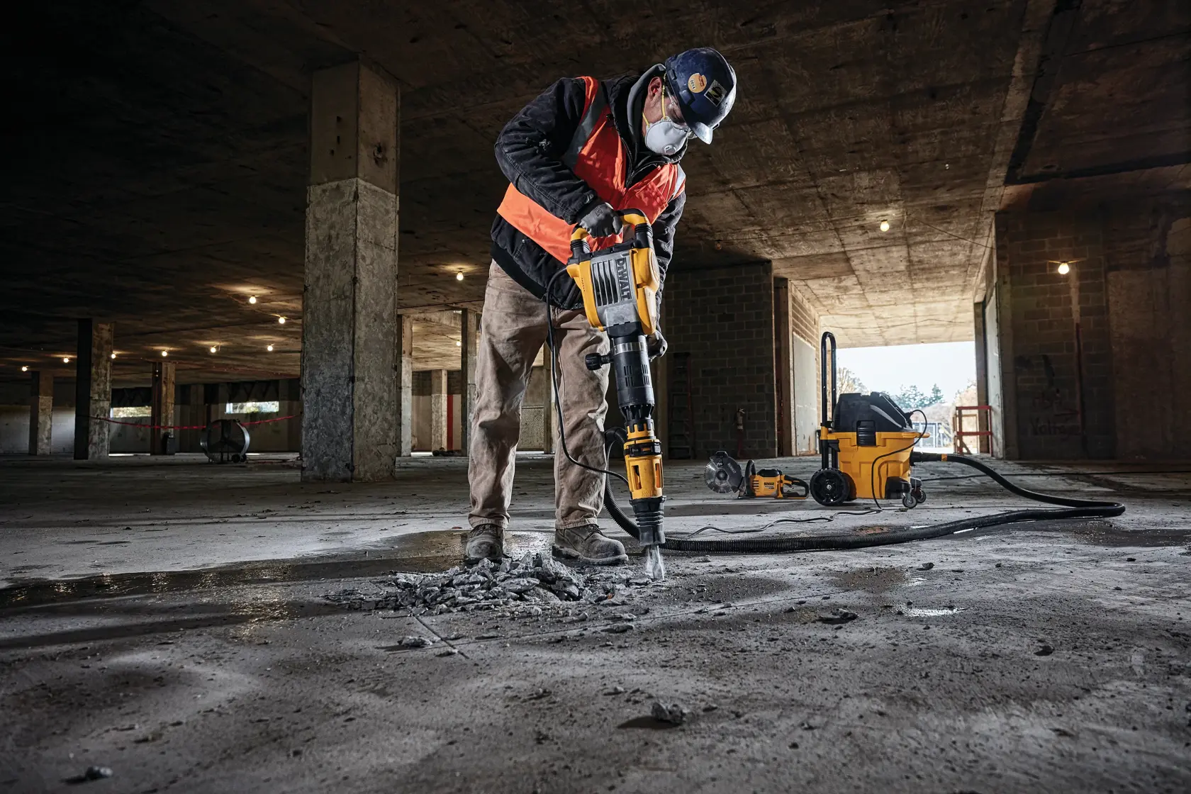 Un trabajador de construcción con equipo de seguridad utiliza un martillo demoledor DEWALT para romper concreto en un edificio sin terminar. Se puede ver un extractor de polvo DEWALT al fondo.