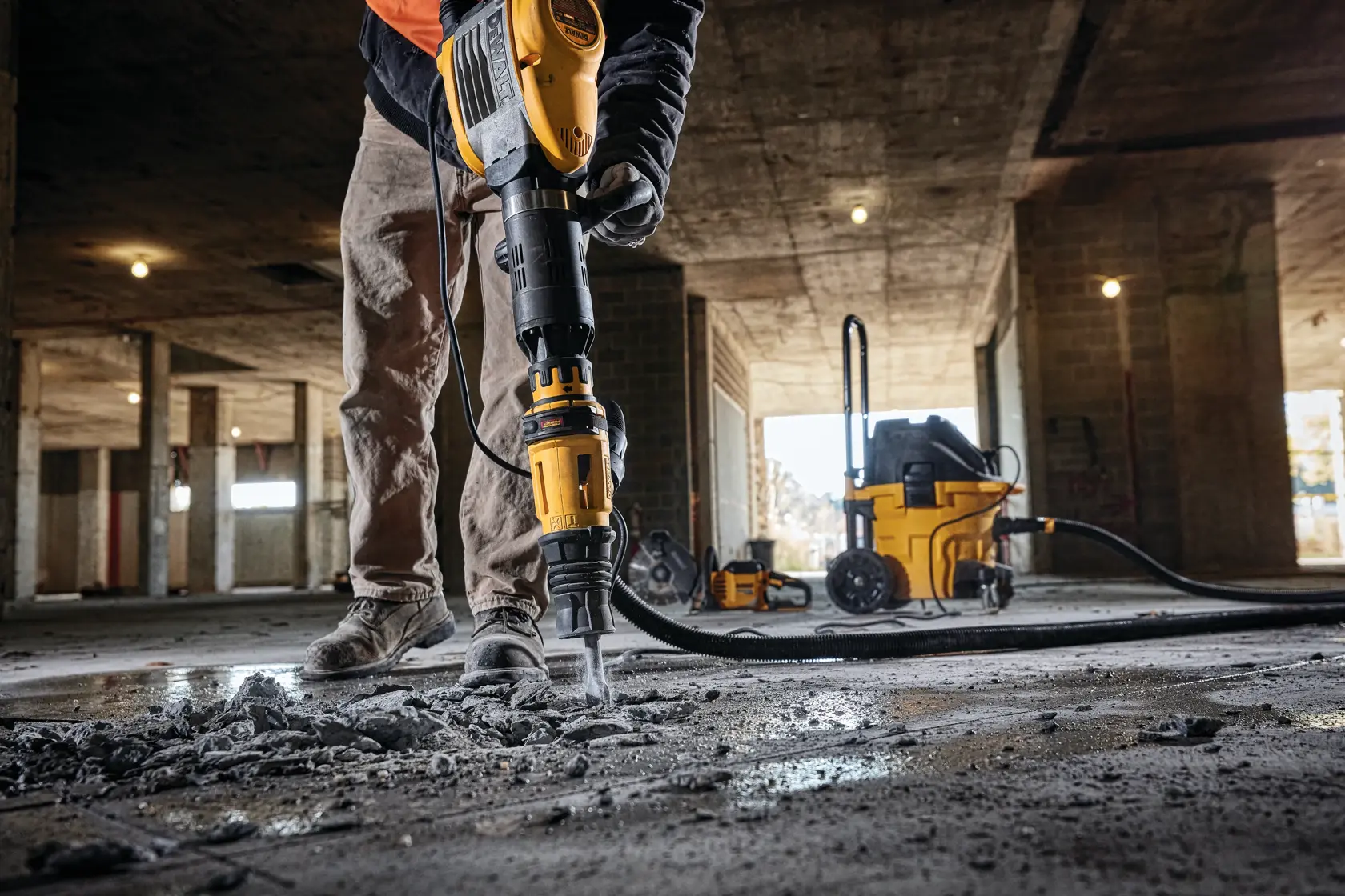 Una persona utiliza un martillo demoledor DEWALT D25951K para romper concreto en un sitio de construcción, con escombros dispersos en el suelo y equipo adicional DEWALT visible en el fondo.