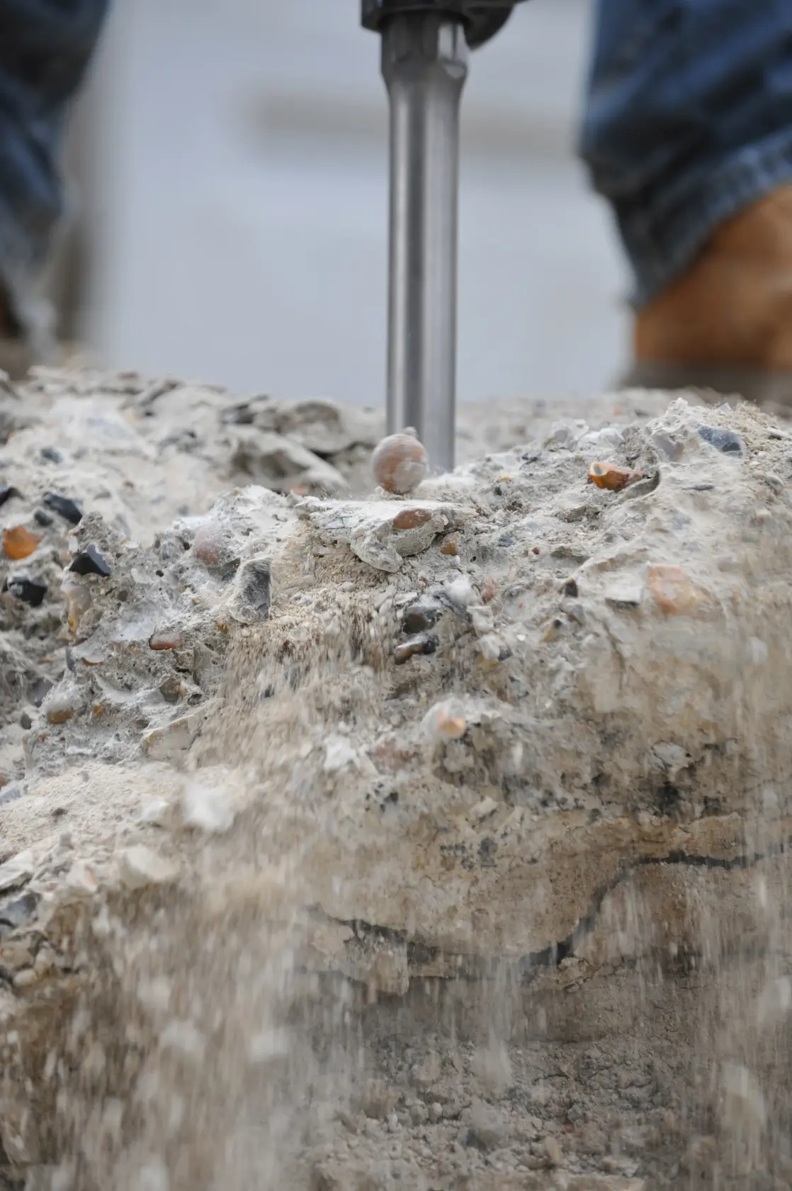 Primer plano de una herramienta de demolición rompiendo concreto, con polvo y escombros cayendo y los pies de una persona visibles al fondo.