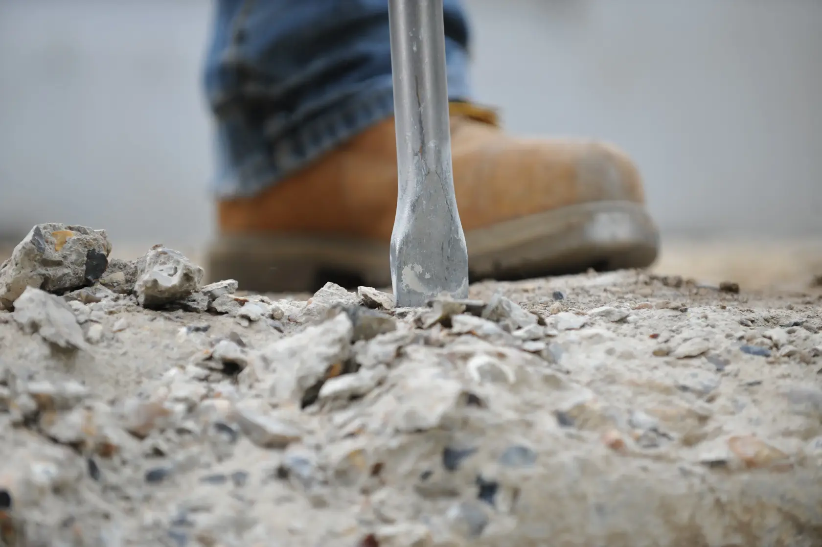 Primer plano de una punta de cincel rompiendo concreto, con un trabajador usando una bota de seguridad marrón en el fondo.