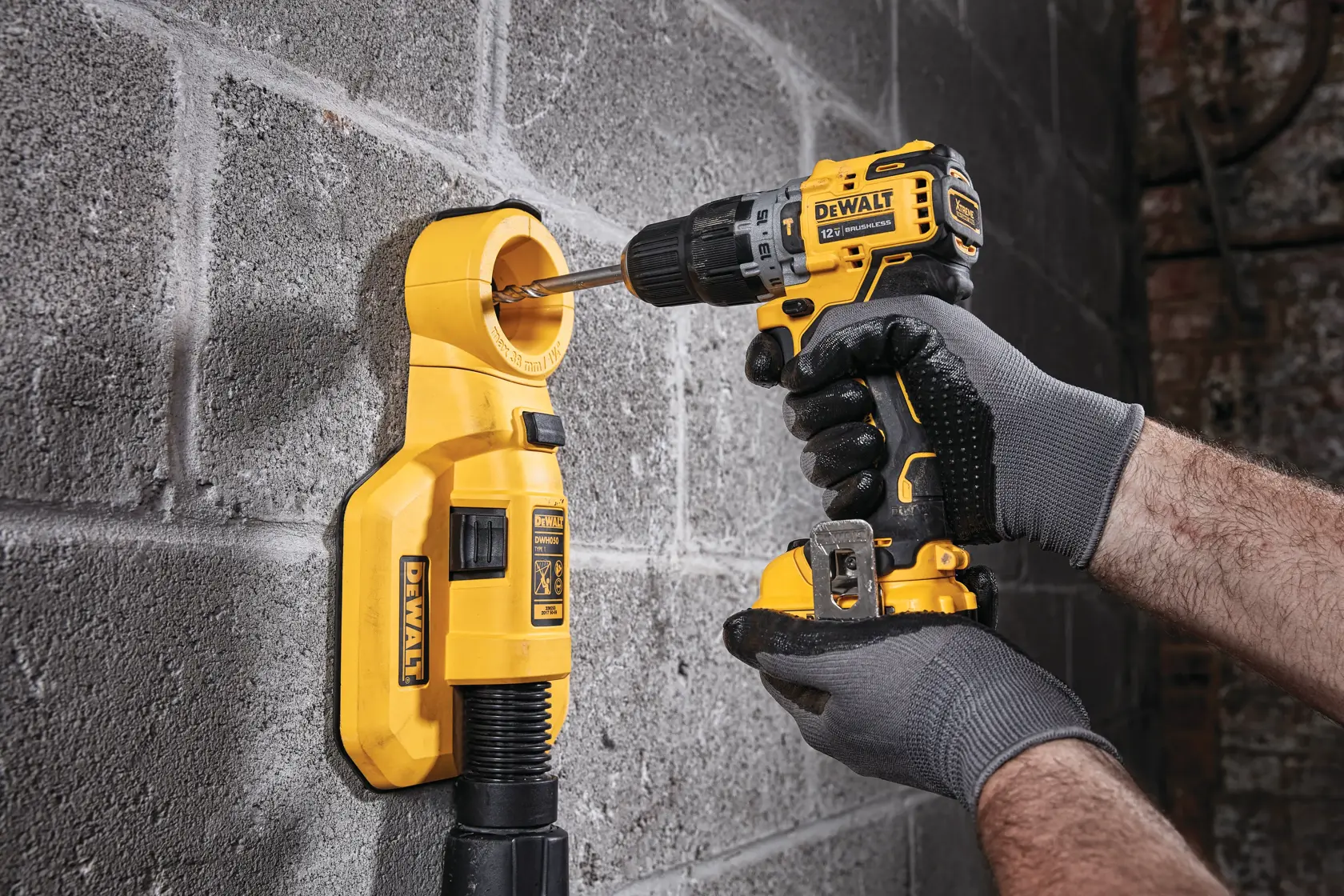 Una persona con guantes está usando un taladro inalámbrico DEWALT para perforar una pared de concreto, mientras un accesorio de aspiradora montado en la pared DEWALT, de color amarillo, recoge el polvo junto a la broca.