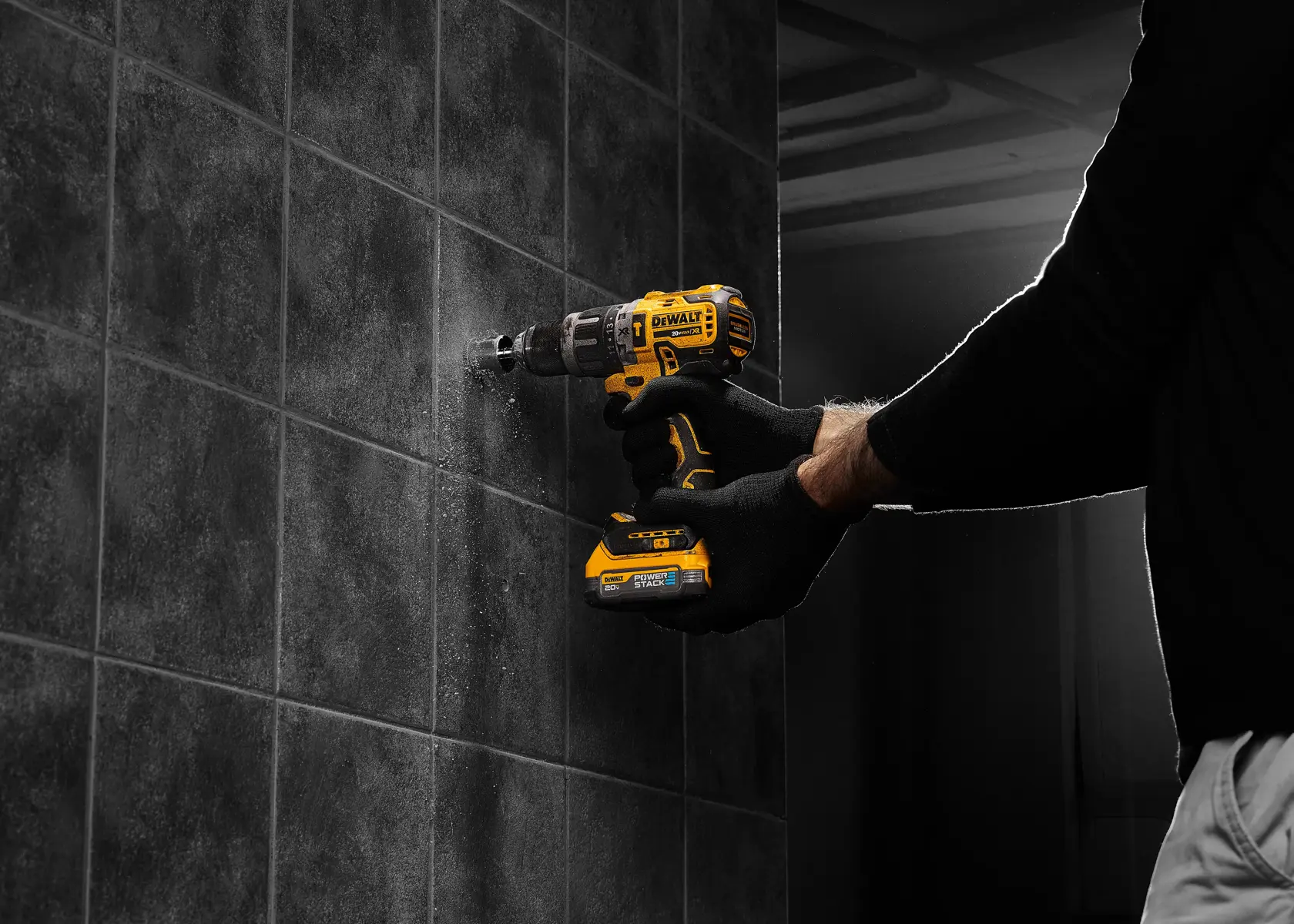 Una persona con guantes negros utiliza un taladro inalámbrico amarillo DEWALT para perforar una pared de baldosas oscuras.