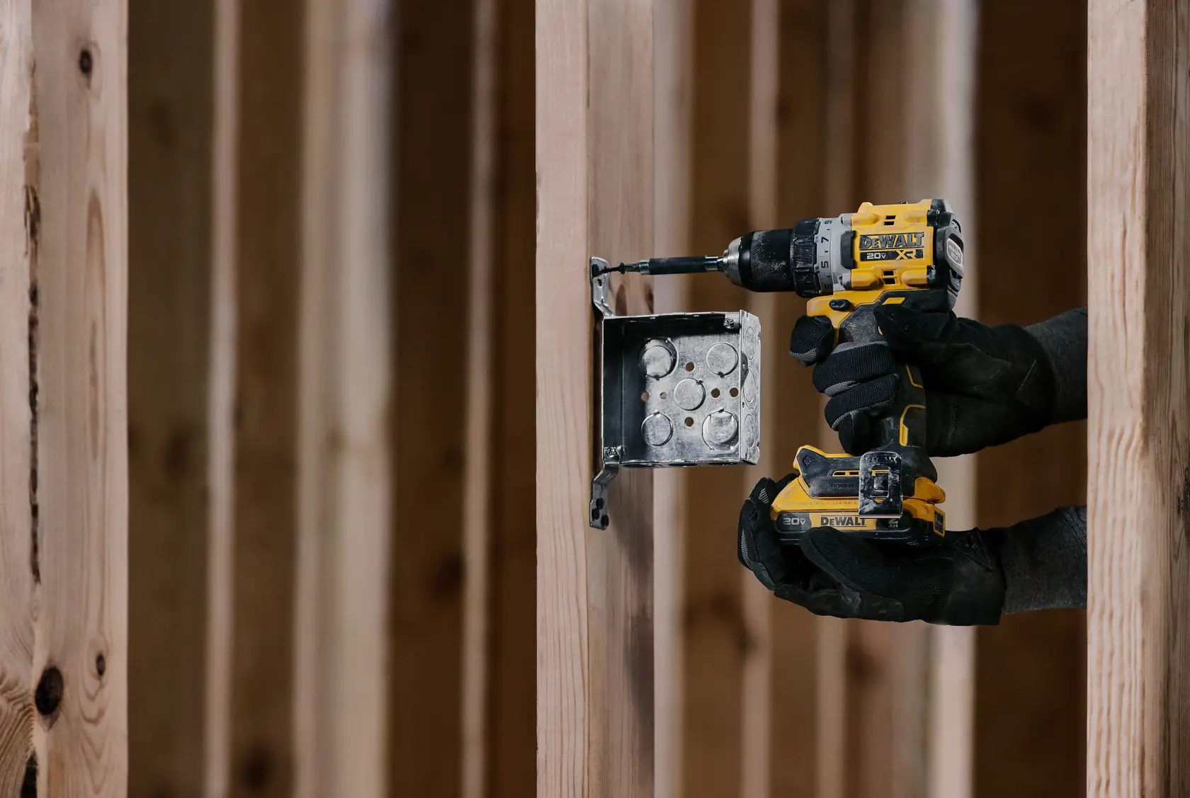 Una persona con guantes utiliza un taladro inalámbrico DEWALT para instalar una caja eléctrica en una estructura de madera en un sitio de construcción.
