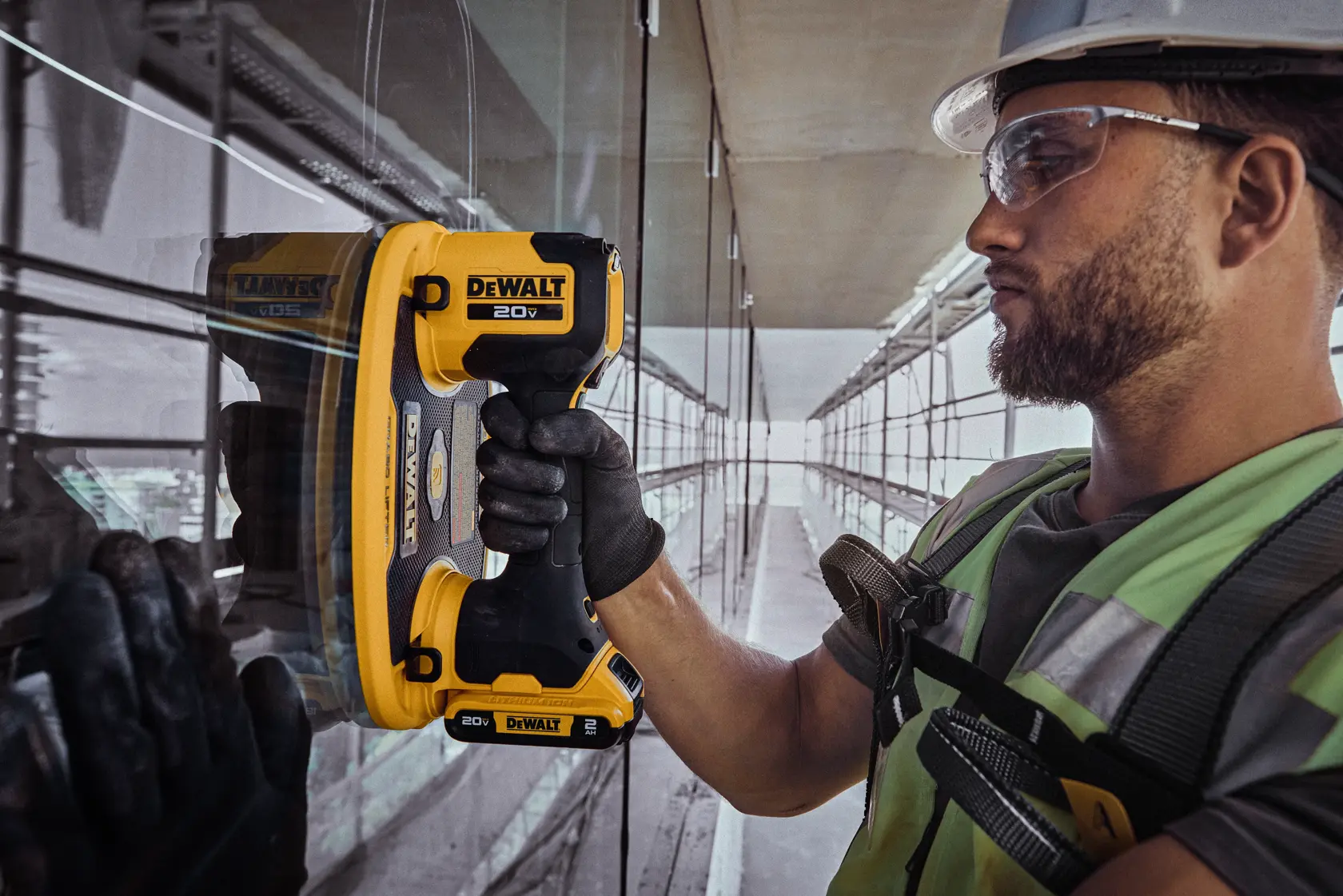 Uma pessoa com equipamento de segurança está usando uma ferramenta portátil DEWALT 20V em uma superfície de vidro em um local de construção, com o rosto desfocado para preservar a privacidade.