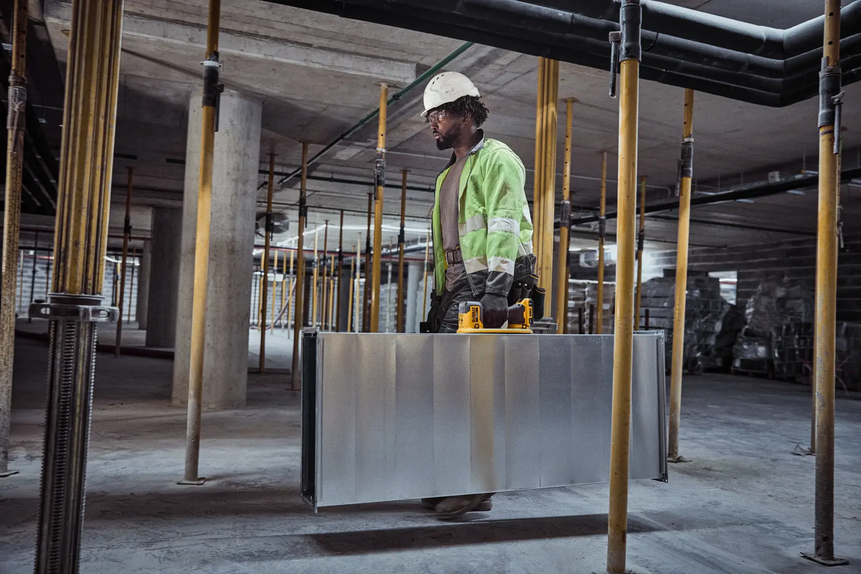 Um trabalhador da construção civil usando jaqueta de alta visibilidade e capacete carrega um grande duto de metal com a ajuda de uma ferramenta elétrica amarela DEWALT dentro de um edifício em construção.