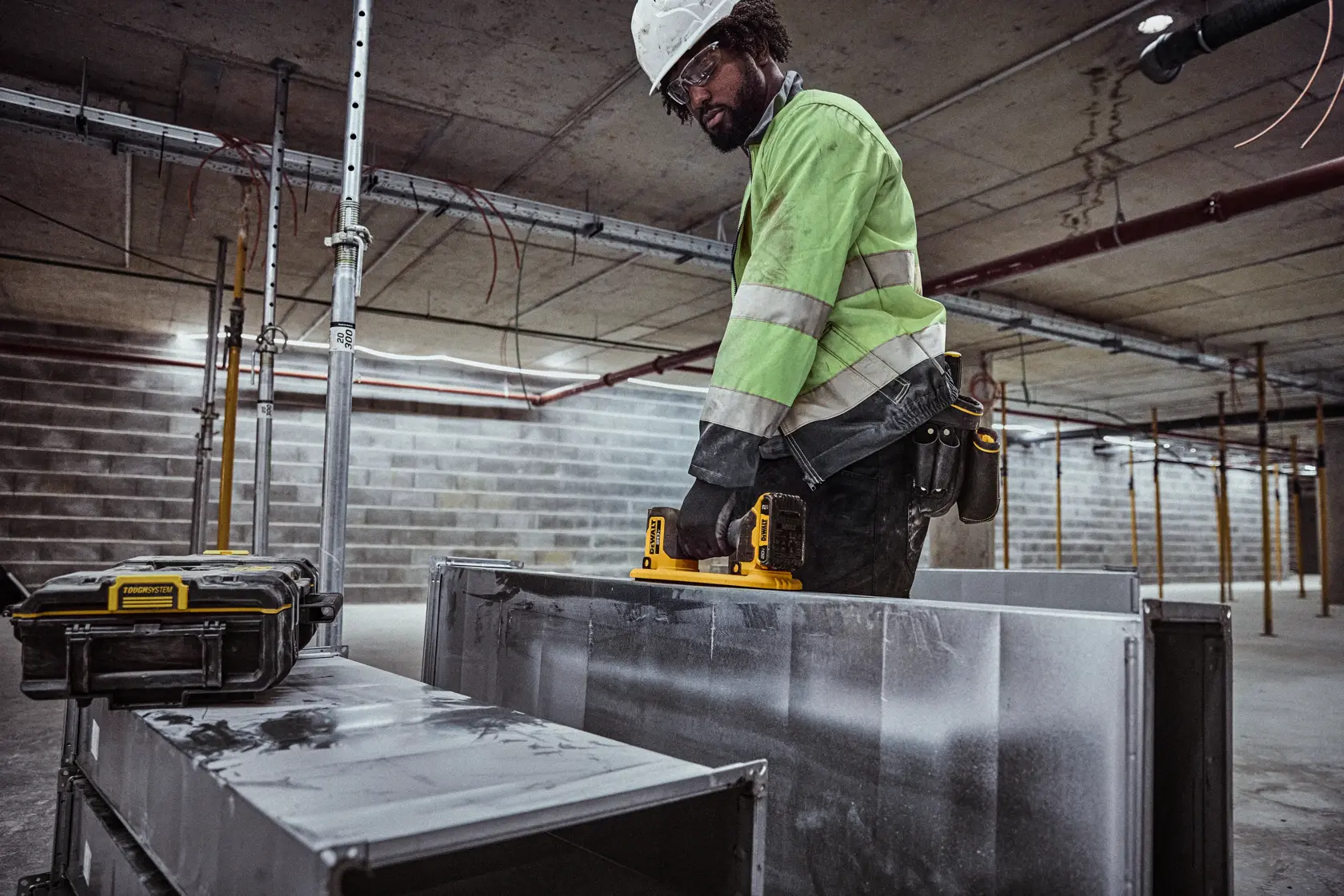 Um trabalhador da construção civil com jaqueta de alta visibilidade utiliza uma câmera de inspeção sem fio DEWALT (DCE592B) para examinar um duto de metal dentro de um prédio em construção. Uma maleta de proteção está próxima.