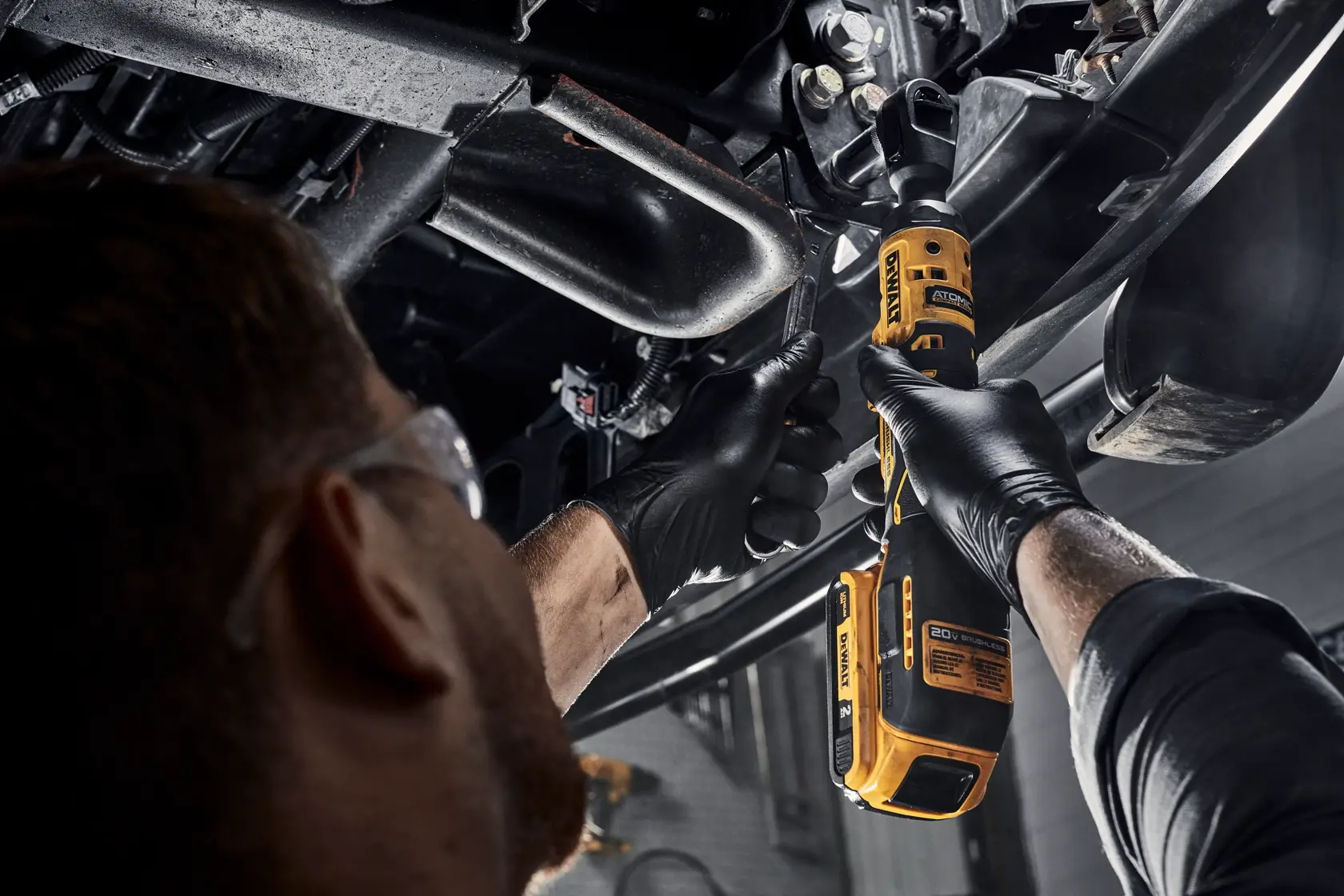 Una persona con gafas de seguridad y guantes negros utiliza una herramienta de carraca inalámbrica amarilla DEWALT para trabajar en piezas mecánicas debajo de un vehículo.