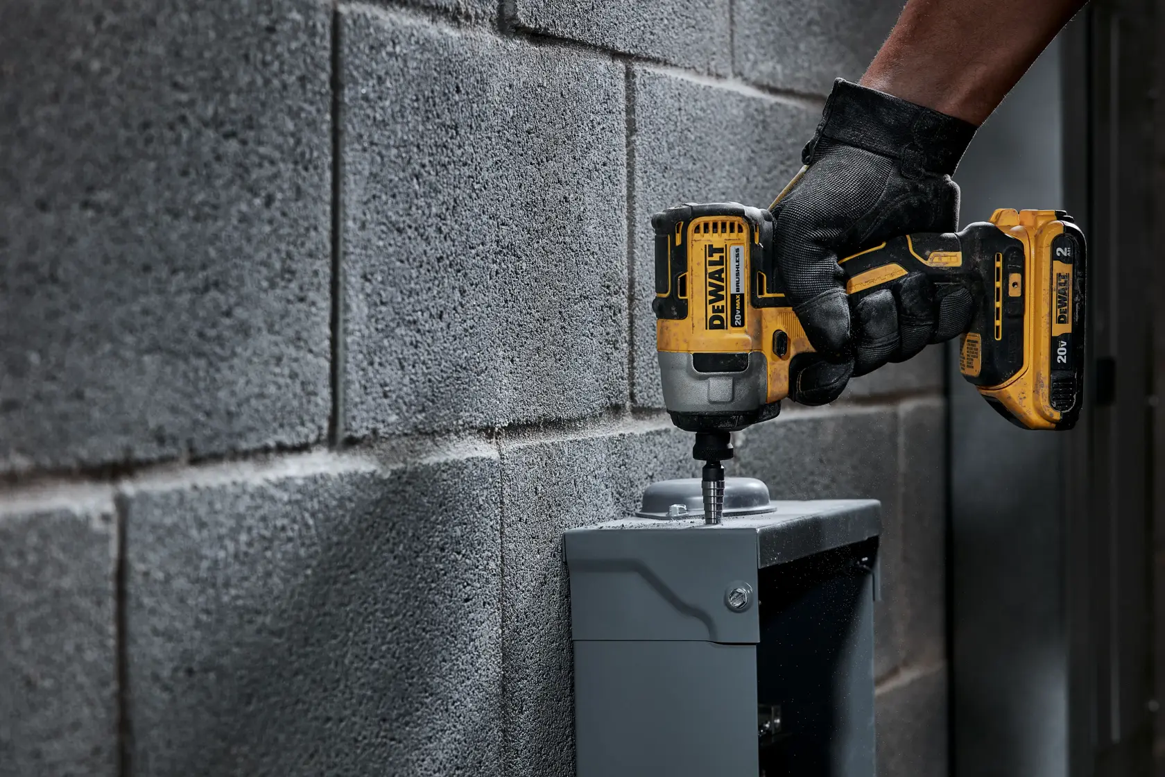 Uma pessoa usando luvas pretas utiliza uma parafusadeira de impacto sem fio amarela DEWALT para fixar um parafuso em uma caixa de metal cinza instalada em uma parede de blocos de concreto.
