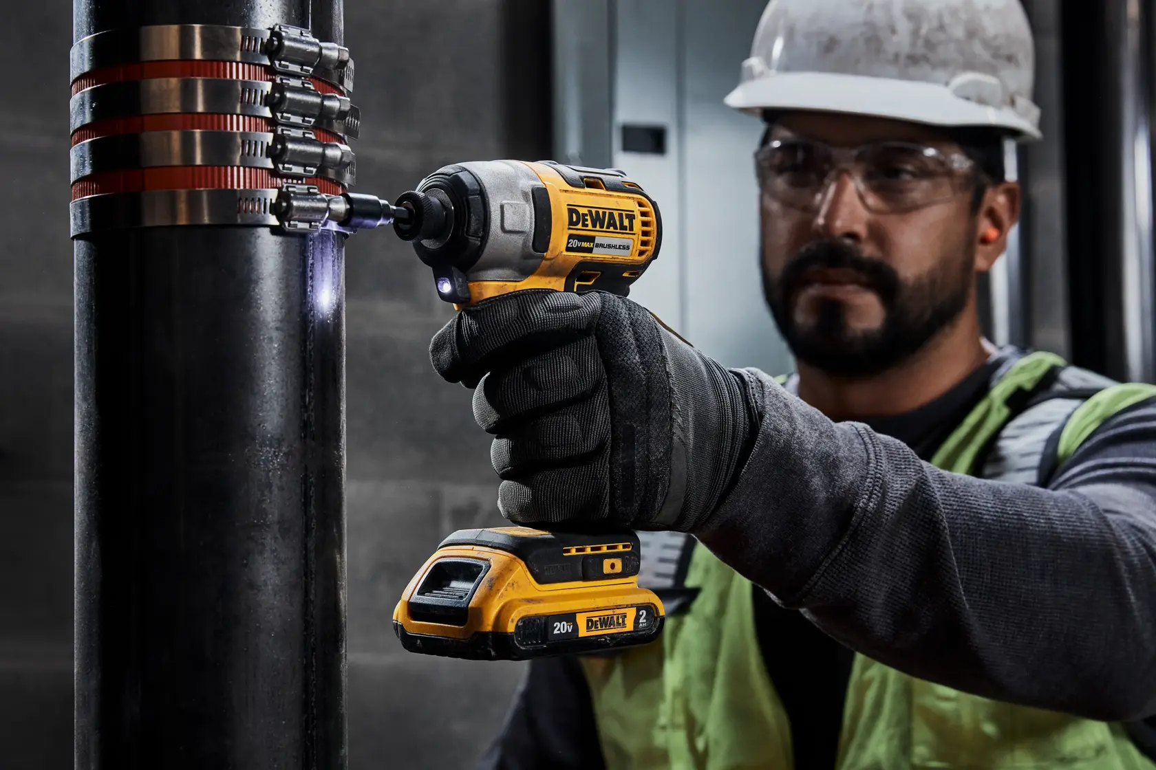Un trabajador con equipo de seguridad utiliza un atornillador de impacto inalámbrico DEWALT para fijar abrazaderas en una tubería grande en interiores. El rostro de la persona está difuminado por privacidad.