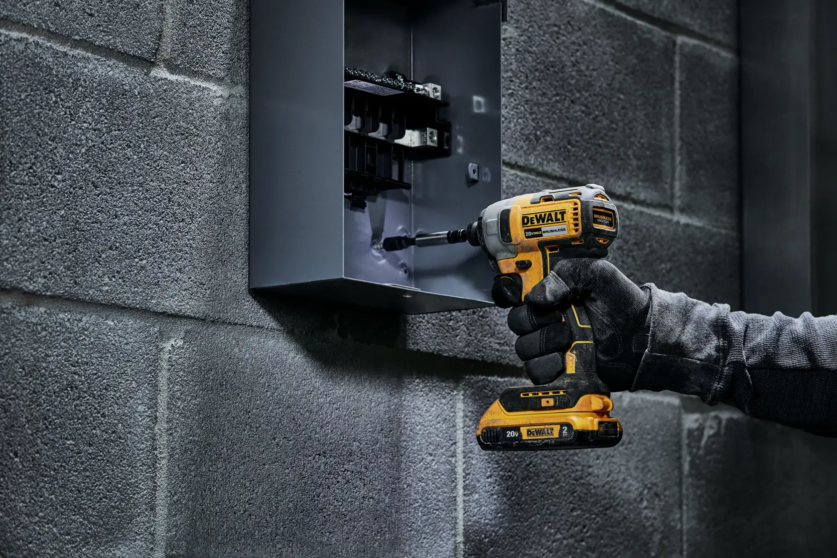 Una persona utiliza un atornillador de impacto inalámbrico DEWALT para trabajar dentro de un panel eléctrico montado en la pared. La persona lleva guantes de protección y opera la herramienta, que es de color amarillo y negro.