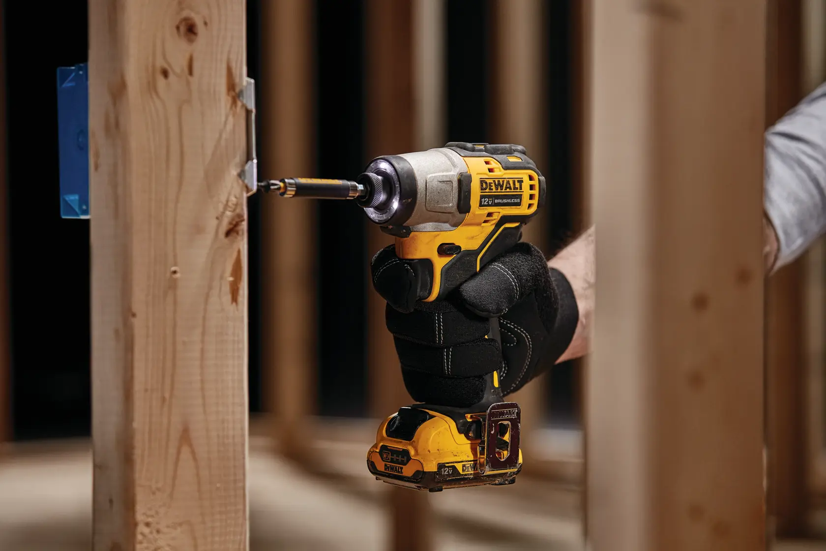 Una persona con guantes de trabajo negros utiliza un atornillador de impacto inalámbrico amarillo y negro DEWALT para perforar un tornillo en un marco de madera.