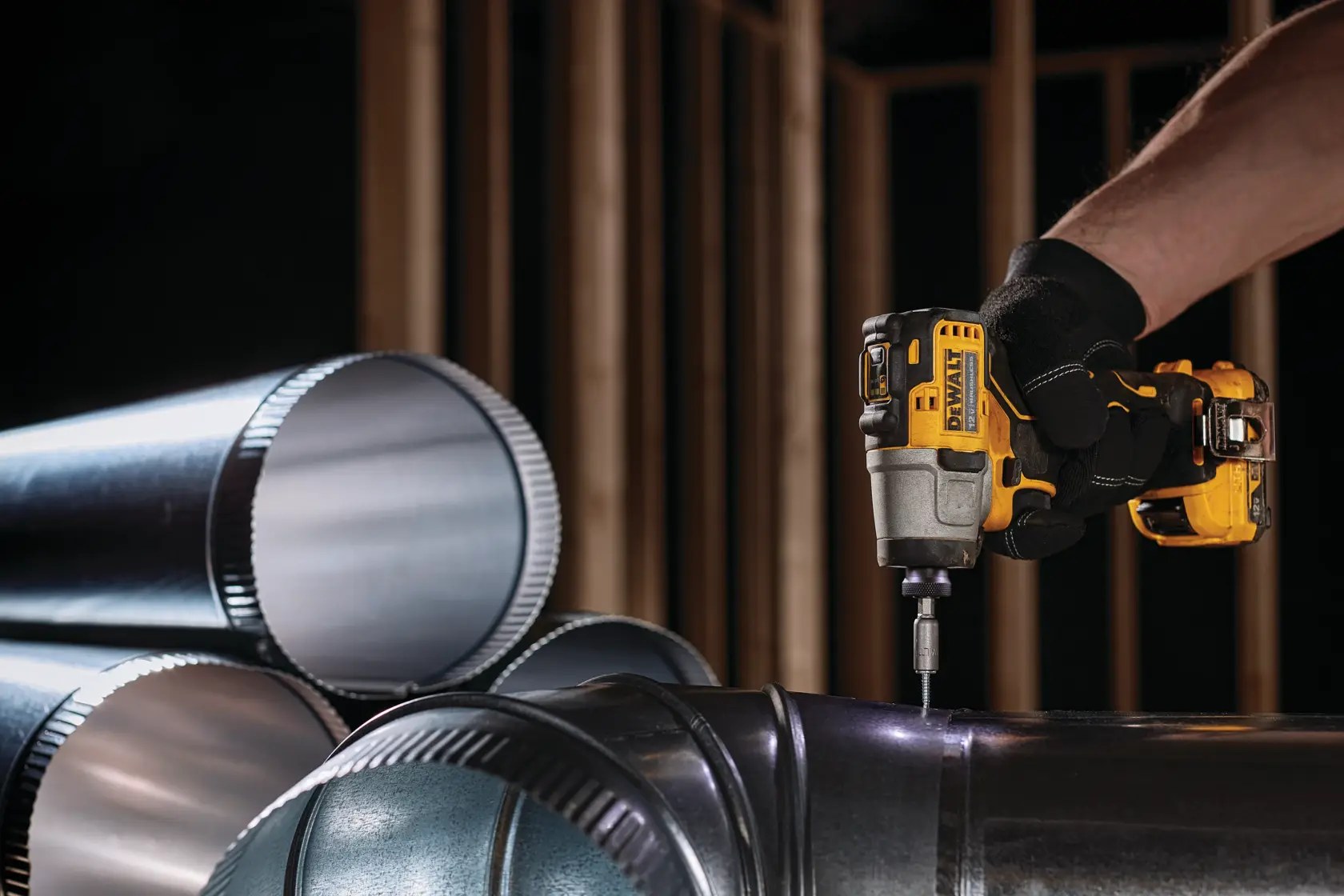 Una persona con guantes negros está usando un destornillador de impacto inalámbrico DEWALT para atornillar un tornillo en tubos de conductos metálicos. Varios tubos de conducto metálicos cilíndricos están apilados en primer plano, y en el fondo se ven estructuras de madera.