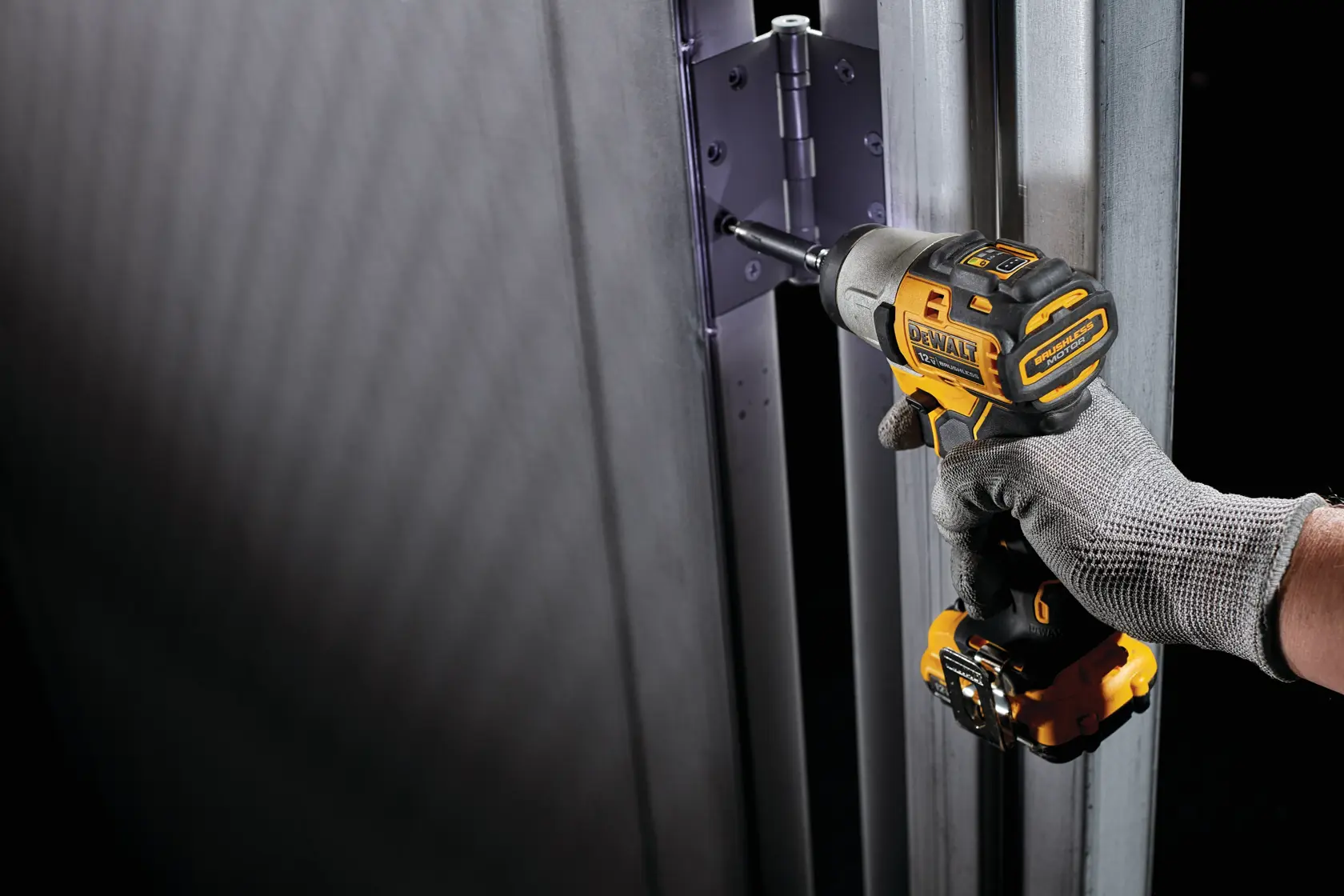 Una mano con un guante gris utiliza un destornillador de impacto inalámbrico DEWALT para ajustar tornillos en una bisagra de metal colocada en un marco metálico.
