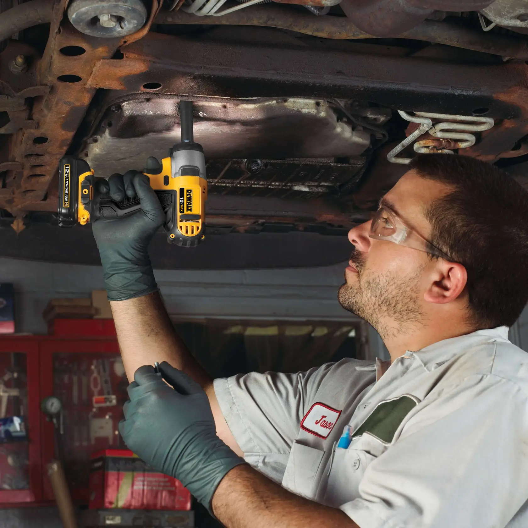 Un mecánico con guantes utiliza un destornillador de impacto inalámbrico DEWALT amarillo y negro debajo de un vehículo, posiblemente para una reparación automotriz. La cara del mecánico está borrosa.
