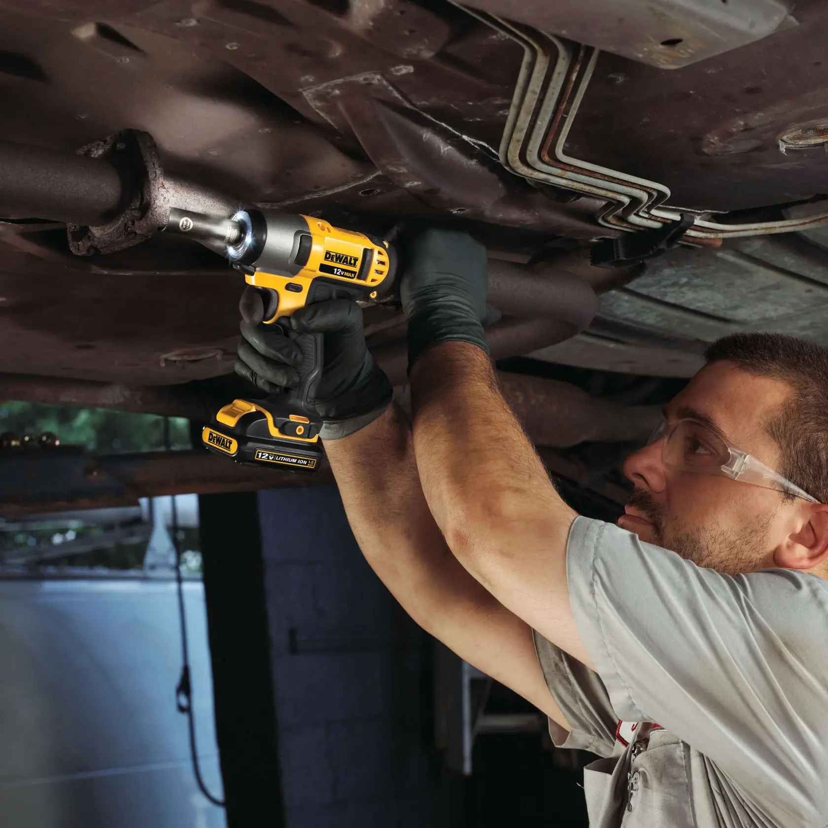 Una persona está utilizando una llave de impacto inalámbrica DEWALT de 12V (modelo DCF813S2) para trabajar en la parte inferior de un vehículo. El individuo lleva guantes y está apretando o aflojando un tornillo en un componente metálico bajo el coche.
