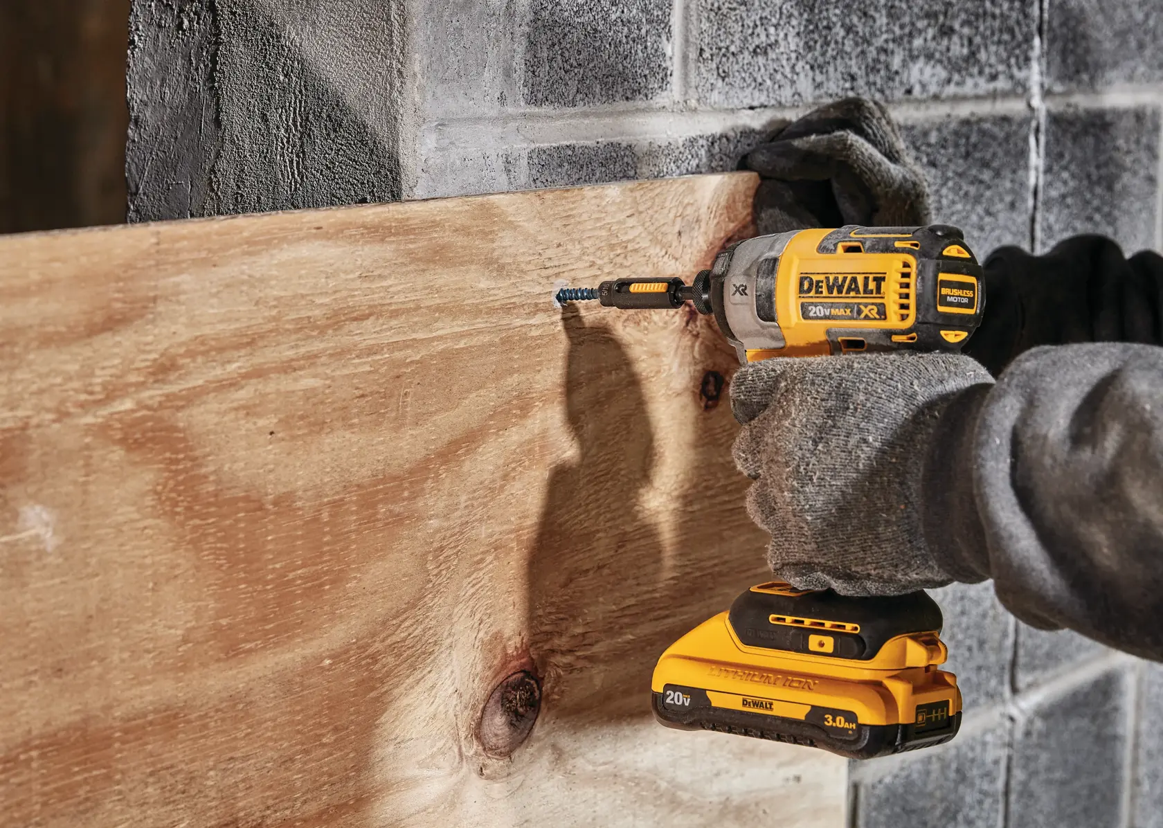 Una persona con guantes utiliza un destornillador de impacto inalámbrico DEWALT 20V Max XR amarillo y negro para atornillar un tornillo en una tabla de madera fijada a una pared de concreto.