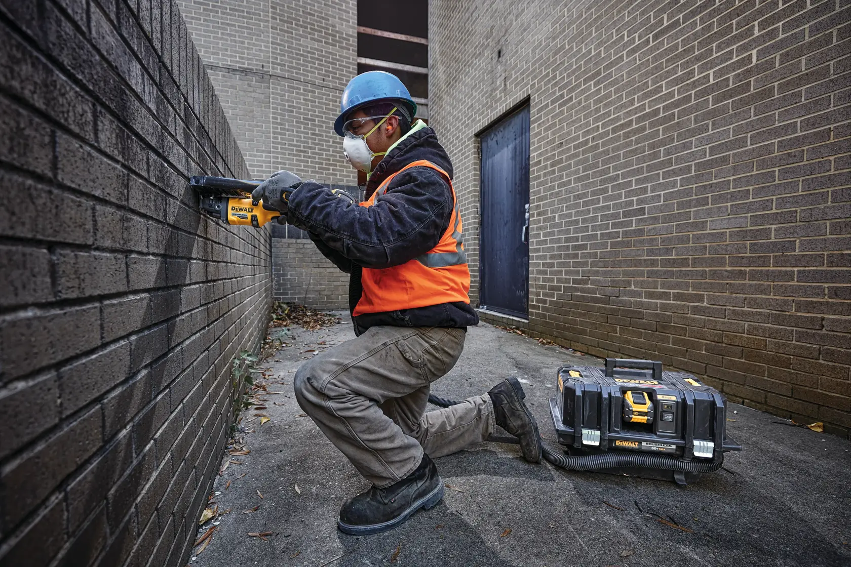 Un trabajador de la construcción con casco azul, chaleco de seguridad naranja y guantes está arrodillado sobre una superficie de concreto junto a una pared de ladrillos, usando una herramienta eléctrica DEWALT. Un estuche de equipo con la marca DEWALT está colocado en el suelo cerca.