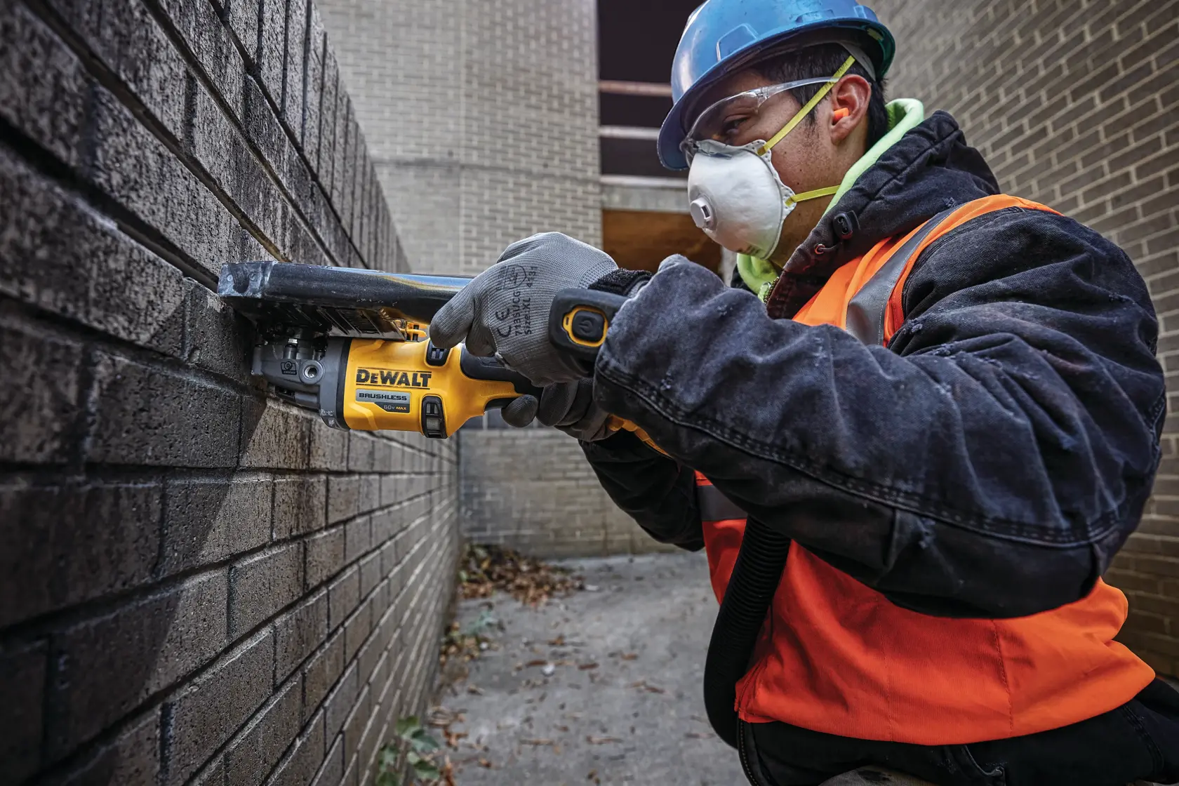 Un trabajador que lleva casco azul, chaleco de seguridad naranja y guantes utiliza una herramienta eléctrica inalámbrica DEWALT para cortar una pared de ladrillo al aire libre. La herramienta tiene un motor sin escobillas y se utiliza para trabajos de construcción o mampostería.