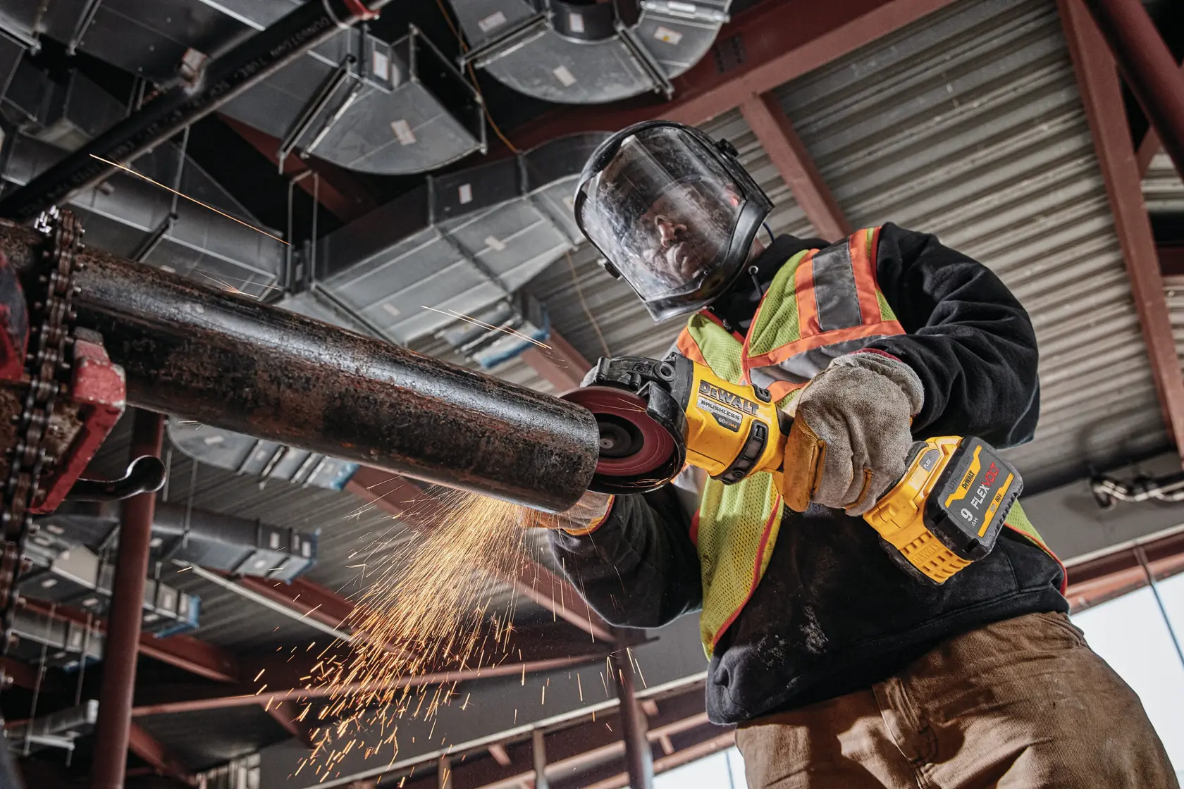 Un trabajador con casco de seguridad, protector facial, guantes y chaleco reflectante utiliza una amoladora angular inalámbrica DEWALT para cortar una tubería de metal, generando chispas, dentro de un edificio industrial.