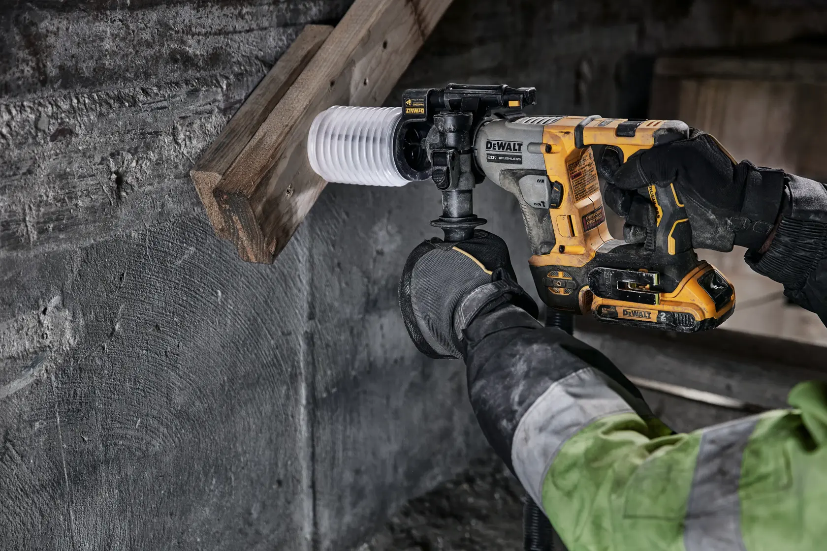 Una persona utiliza un taladro rotativo inalámbrico DEWALT para perforar un agujero a través de una viga de madera y una pared de concreto. El usuario lleva guantes y una chaqueta de trabajo.