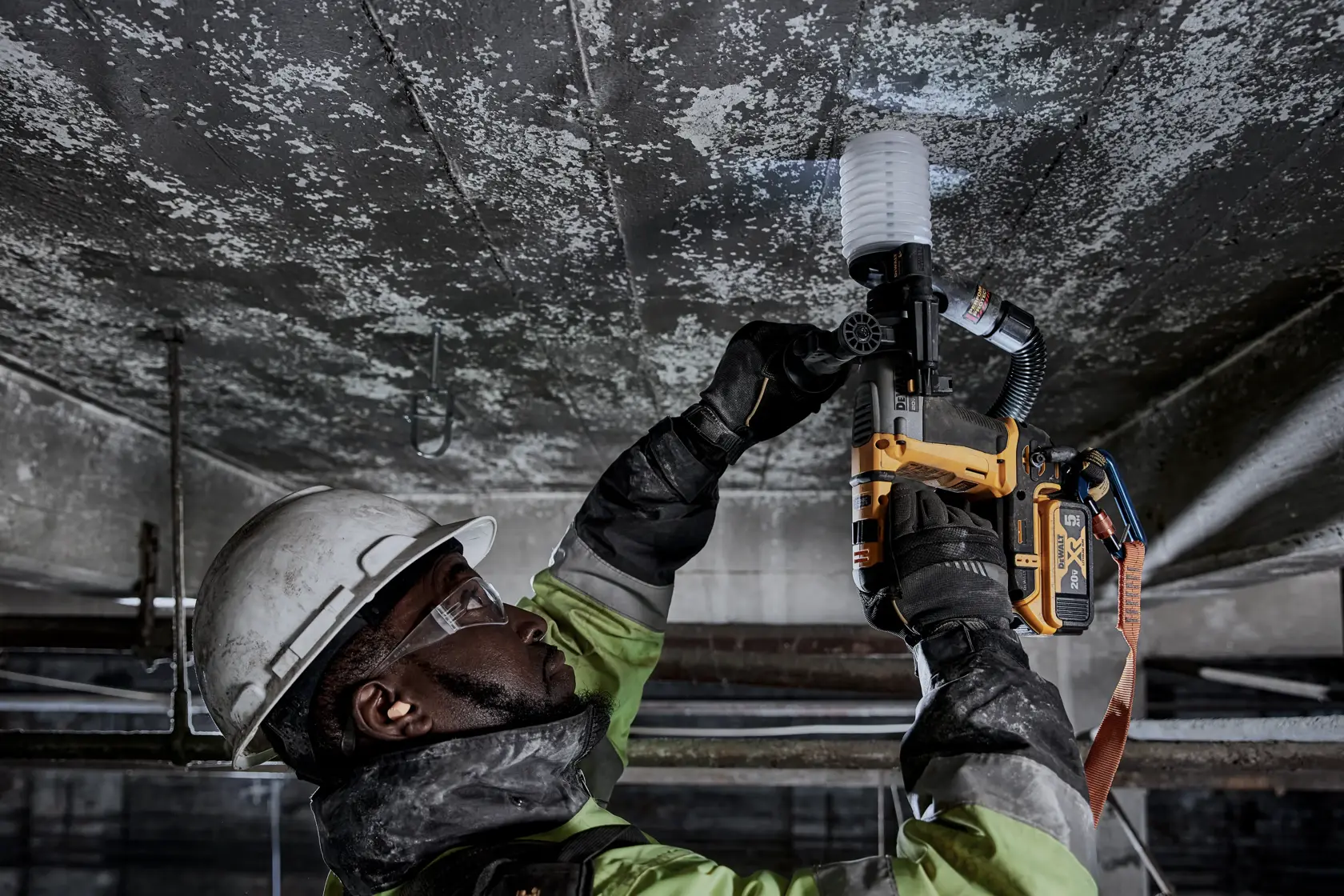 Un trabajador equipado con casco, guantes y gafas protectoras utiliza una herramienta eléctrica amarilla DEWALT para perforar un techo de concreto en un entorno industrial.