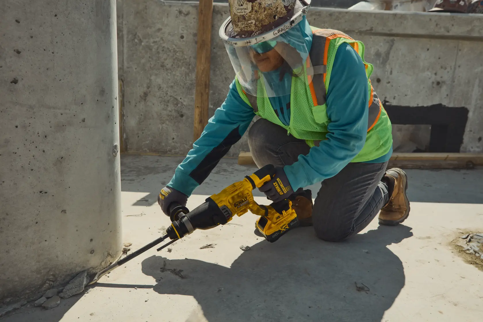 Um trabalhador da construção civil com equipamentos de segurança usa um martelete rotativo amarelo DEWALT (modelo DCH263B) para quebrar concreto na base de um pilar em um canteiro de obras.