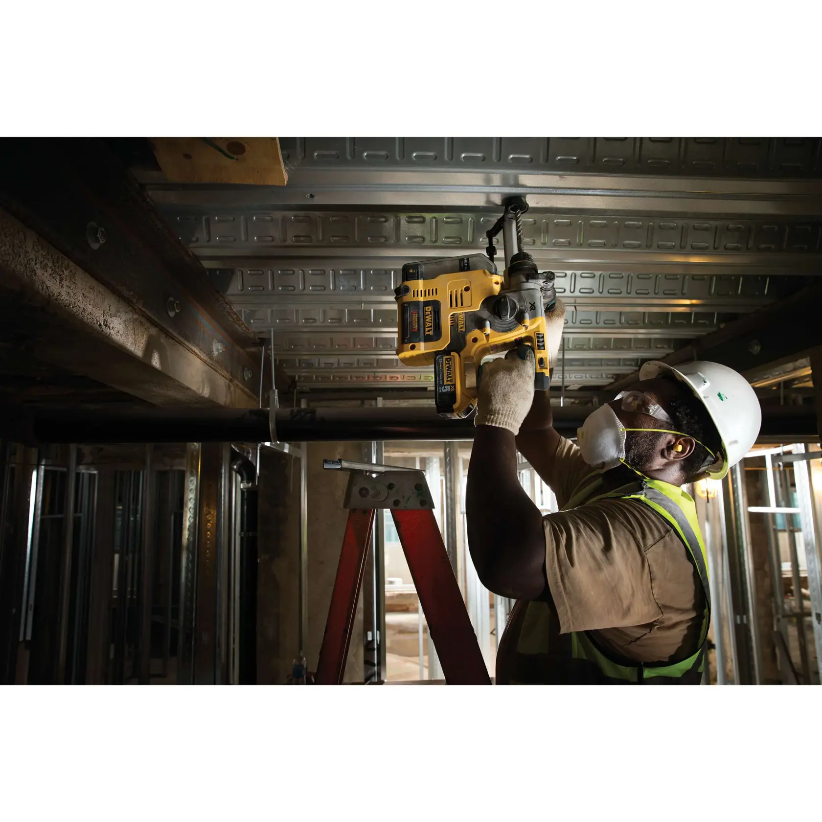 Un trabajador de construcción con casco de seguridad, chaleco reflectante, guantes y mascarilla utiliza un martillo rotativo amarillo DEWALT (modelo DCH273P2) para perforar un techo metálico, de pie sobre una escalera roja dentro de un edificio industrial sin terminar.
