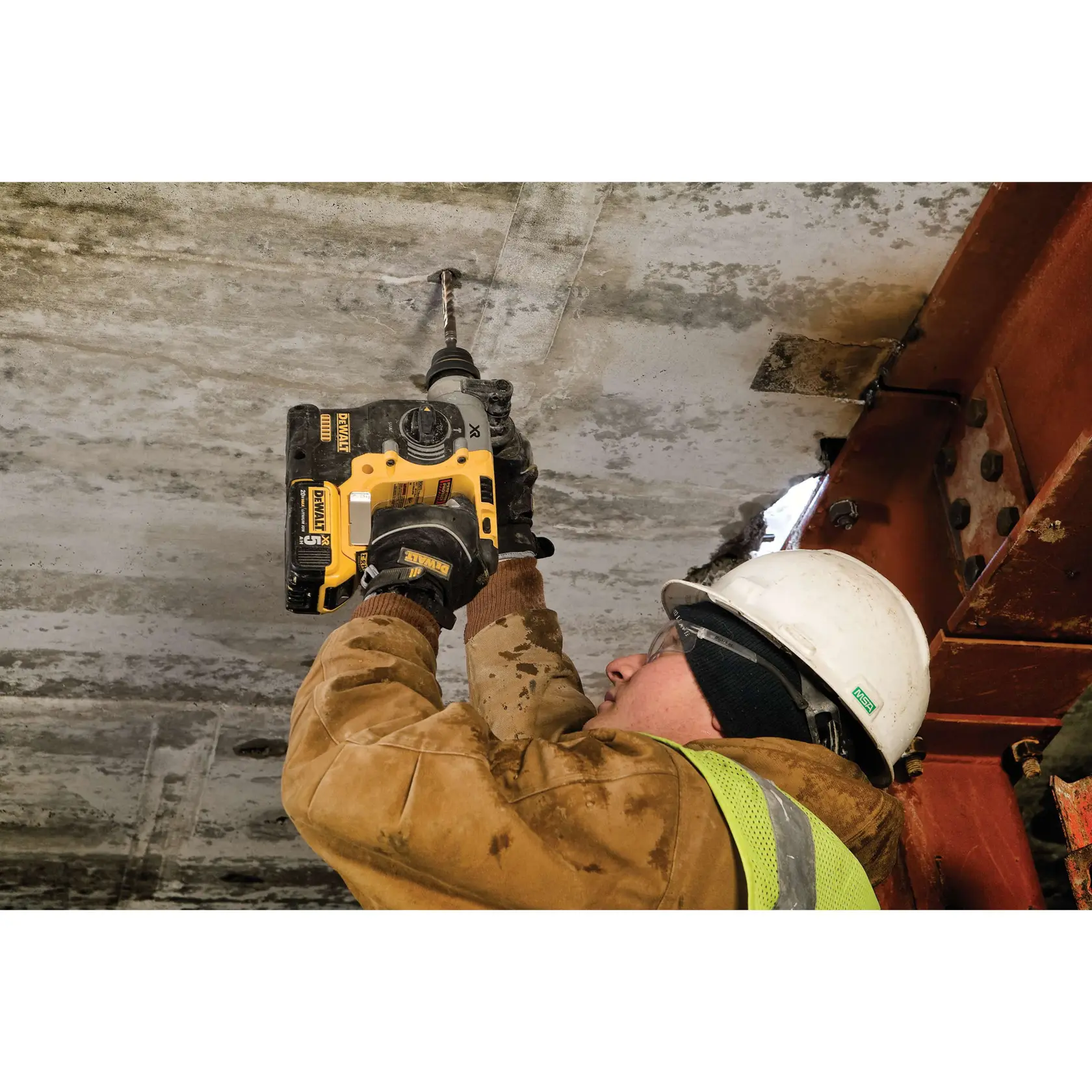 Un trabajador de la construcción con casco de seguridad y chaleco utiliza un martillo rotativo DEWALT DCH273P2 para perforar un techo de concreto. El trabajador lleva una chaqueta protectora marrón y guantes, y está trabajando en una estructura de edificio.