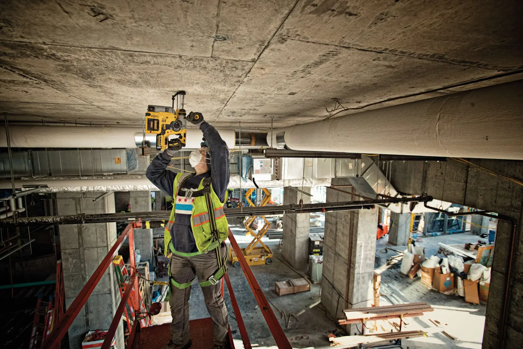 Un trabajador de la construcción con un chaleco de seguridad y mascarilla utiliza una herramienta eléctrica DEWALT para perforar un techo de concreto en una obra interior. El trabajador está de pie sobre una plataforma elevada rodeada de equipos industriales y materiales de construcción.