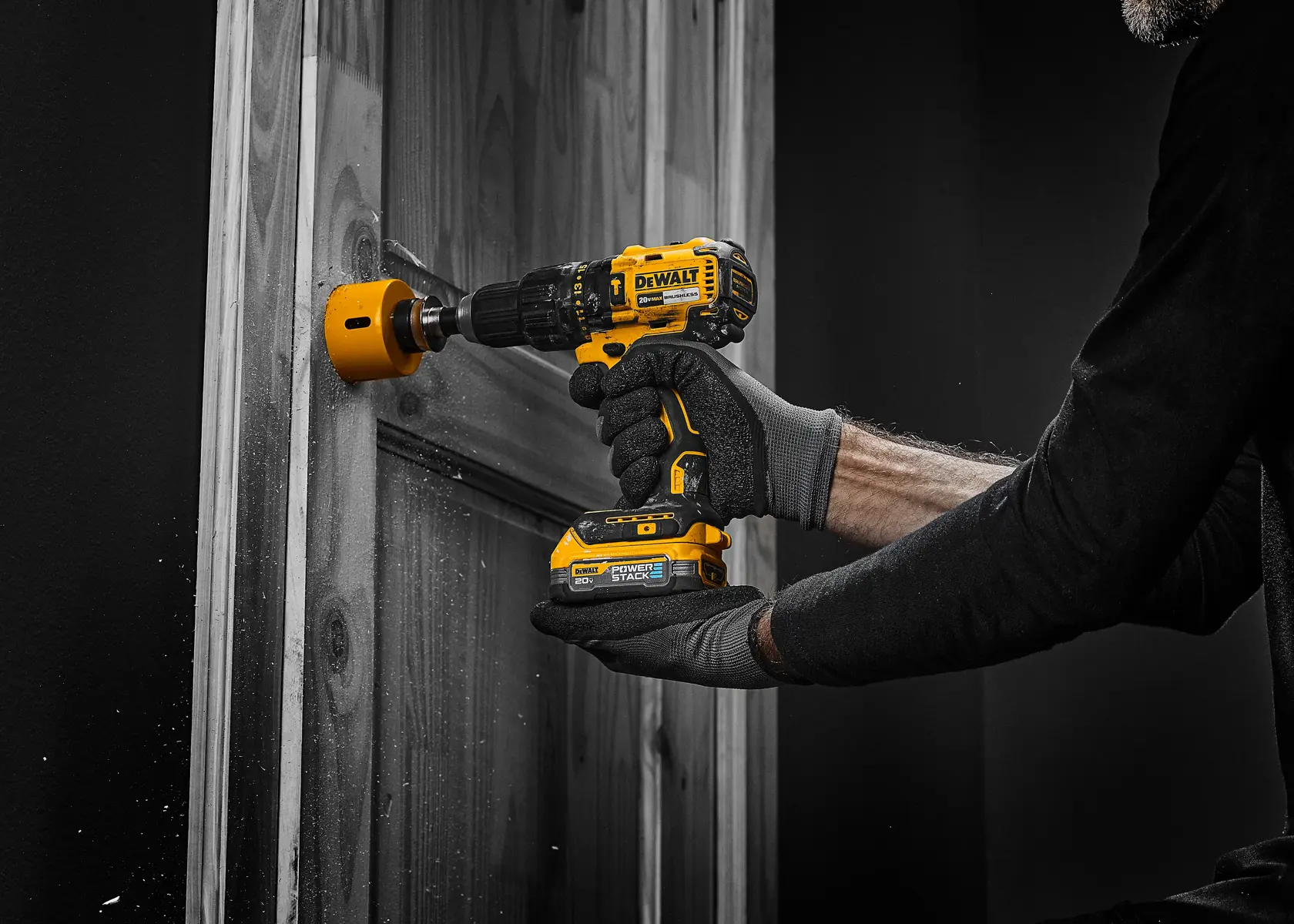 Una persona con guantes está usando un taladro inalámbrico amarillo DEWALT con una broca de corona para perforar un agujero en una puerta de madera.