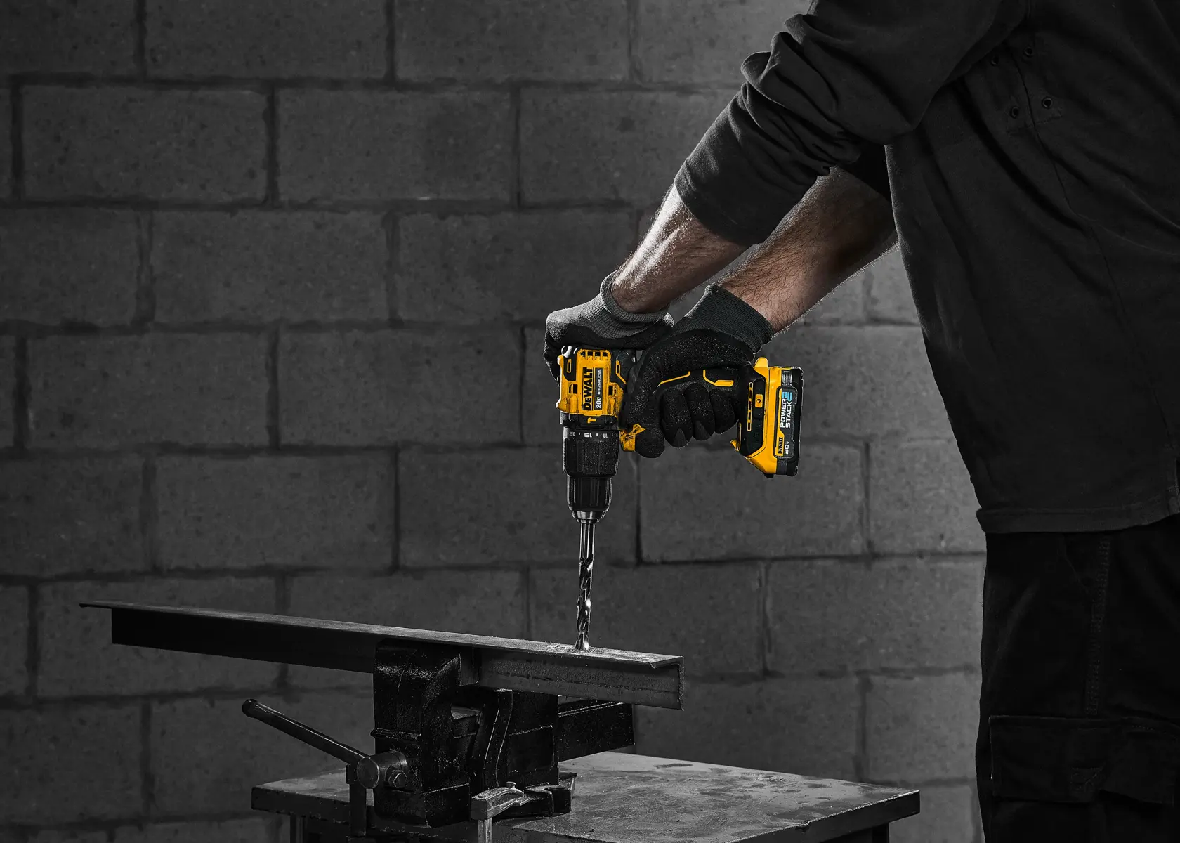 Una persona vestida de negro y con guantes está utilizando un taladro inalámbrico amarillo DEWALT para perforar una barra de metal sujetada a un banco de trabajo. El fondo es una pared de bloques de concreto.
