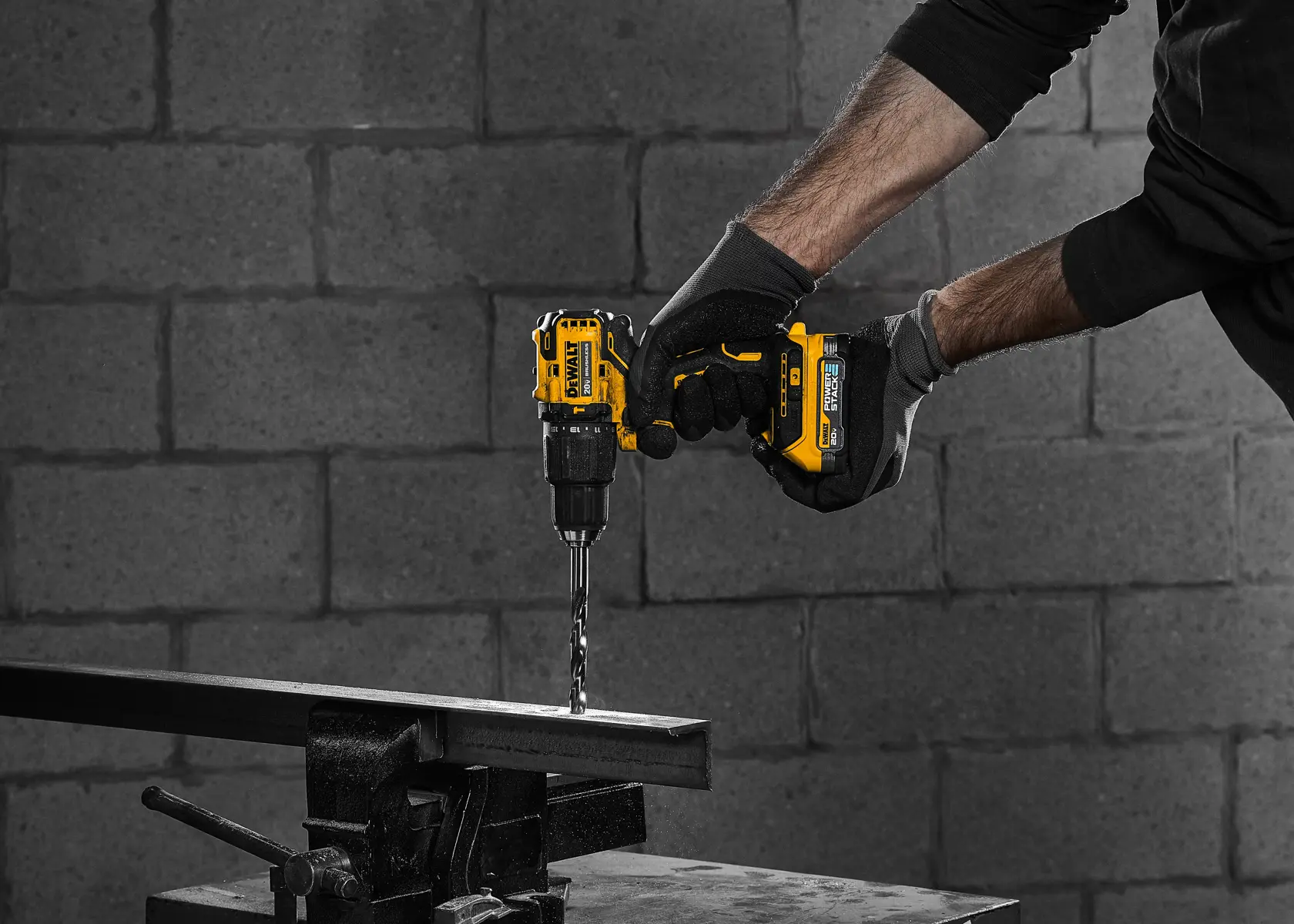 Una persona que lleva guantes está usando un taladro inalámbrico amarillo DEWALT para perforar una barra de metal. La escena ocurre en interiores, con una pared de ladrillos grises de fondo.