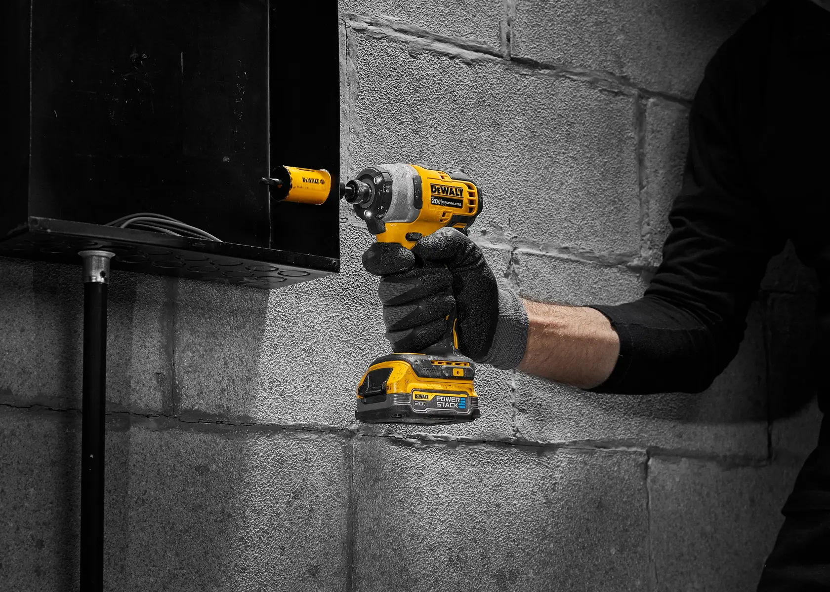 Una persona con ropa negra y guantes utiliza un taladro inalámbrico amarillo DEWALT con una broca de sierra para perforar una caja de metal montada en una pared de concreto.