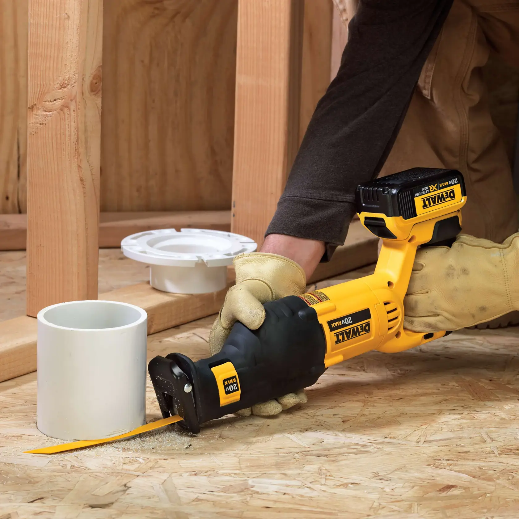 Uma pessoa usando luvas de trabalho utiliza uma serra recíproca sem fio DEWALT 20V Max para cortar um tubo de PVC branco em um piso de madeira, com estrutura de madeira e componentes de encanamento visíveis ao fundo.