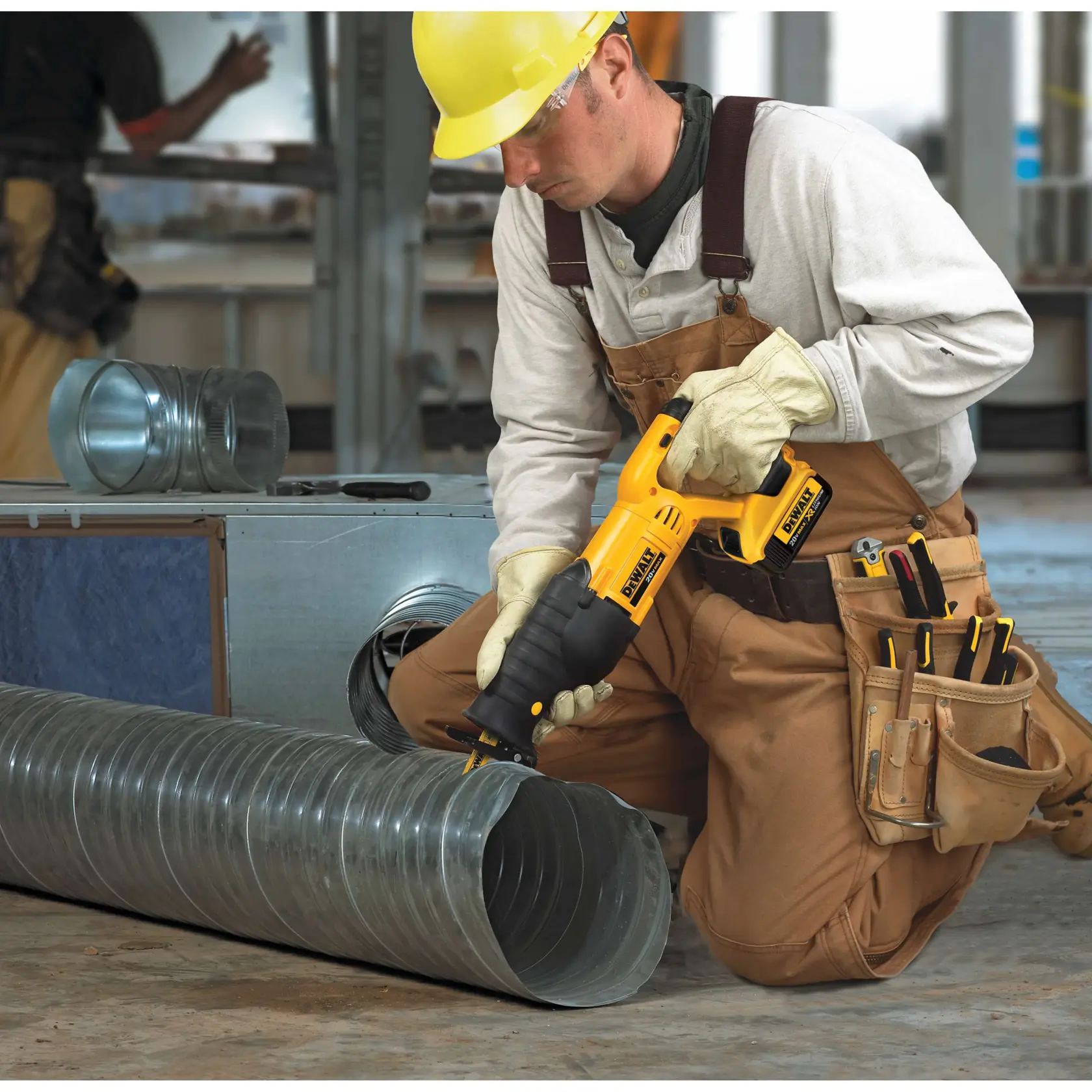 Um trabalhador da construção civil com macacão marrom, luvas e capacete amarelo utiliza uma serra recíproca sem fio DEWALT (modelo DCS380B) para cortar um tubo metálico em um local de trabalho industrial. O trabalhador tem ferramentas em um bolso e está ajoelhado cercado por componentes metálicos.