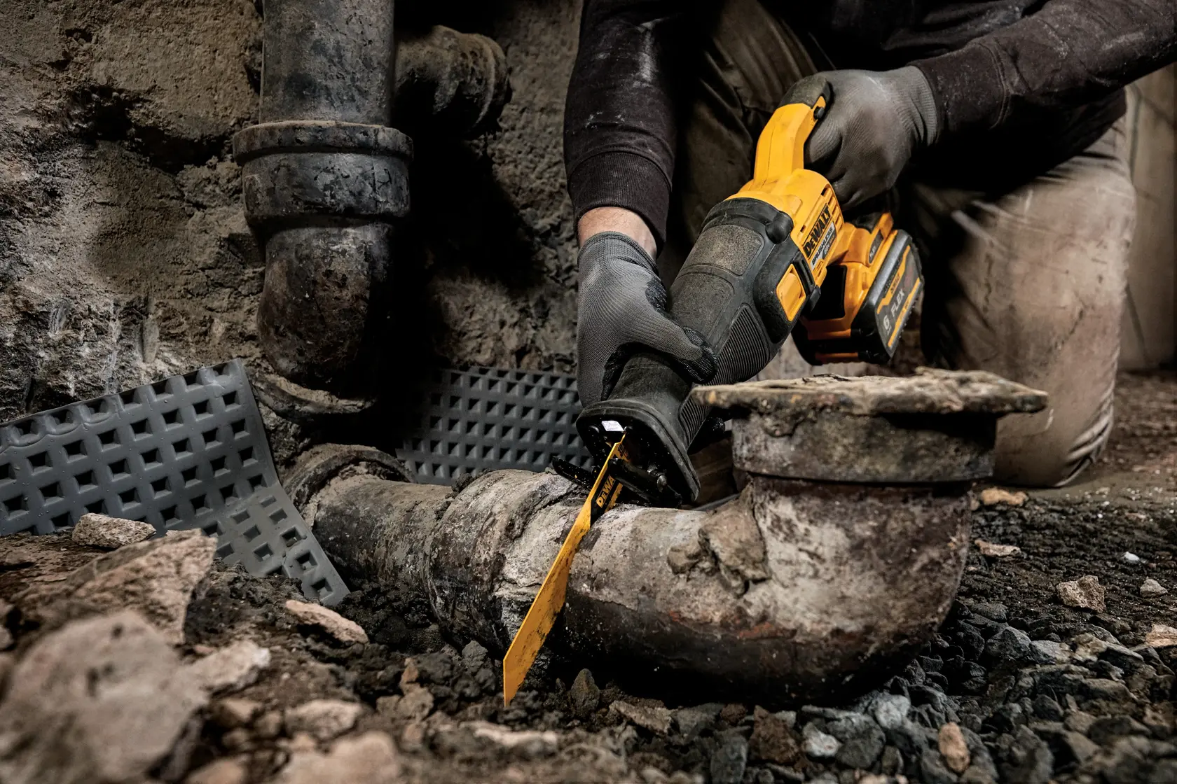 Una persona con guantes está usando una sierra recíproca DEWALT para cortar una tubería de metal vieja en un entorno industrial áspero.