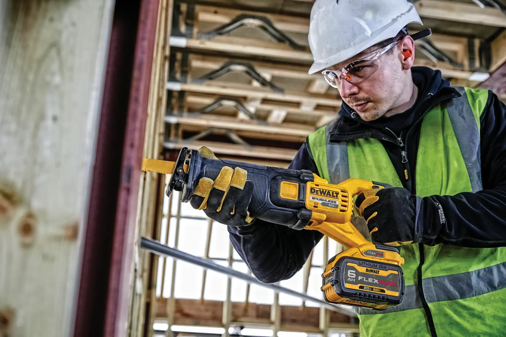 Una persona con guantes de seguridad, chaleco reflectante y casco utiliza una sierra recíproca inalámbrica DEWALT en una obra de construcción. La sierra cuenta con una batería FLEXVOLT y se sostiene horizontalmente mientras corta una viga de madera.