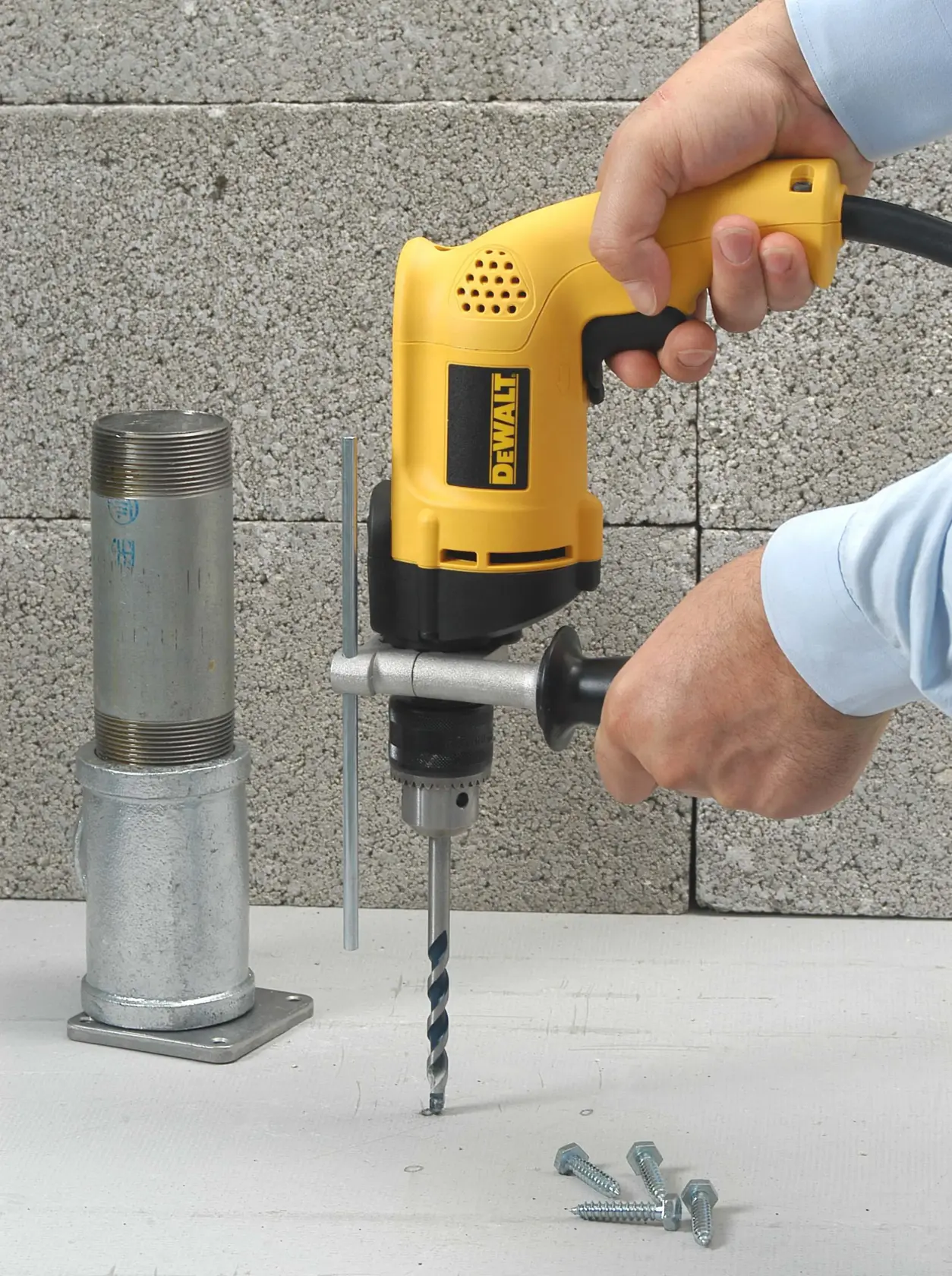 Una persona está usando un taladro eléctrico amarillo DEWALT con mango lateral para perforar una superficie junto a una base de tubería metálica. Hay varios tornillos sobre la superficie de trabajo blanca y bloques de hormigón al fondo.