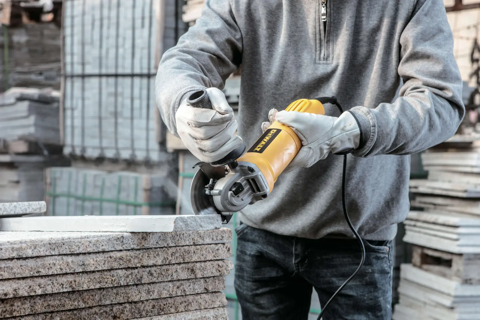 Una persona con guantes utiliza una amoladora angular amarilla DEWALT para cortar losas de piedra en un entorno industrial o de construcción.