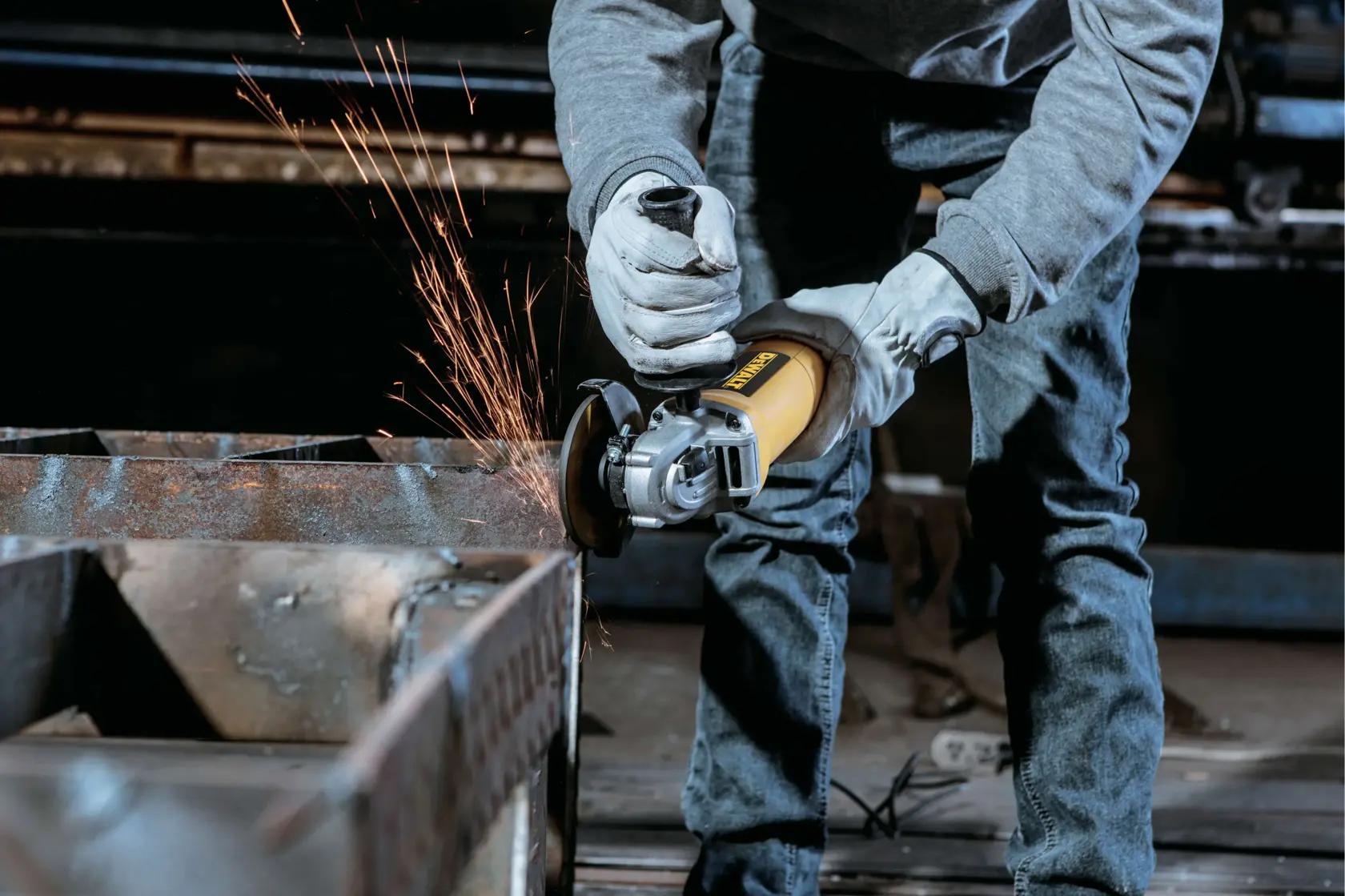 Una persona con guantes y jeans está usando una amoladora angular amarilla DEWALT, generando chispas mientras corta o lija una superficie metálica en un entorno de taller.