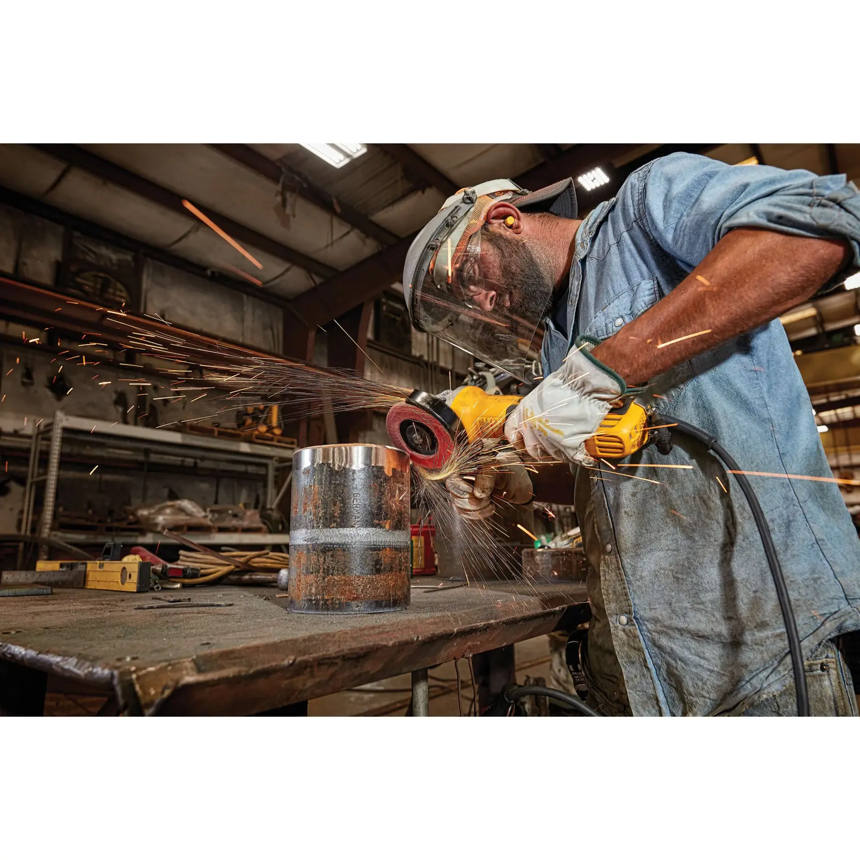 Un trabajador con equipo de protección, incluyendo careta, tapones para los oídos, guantes y camisa de mezclilla, usa una amoladora angular amarilla DEWALT para lijar un cilindro de metal sobre una mesa de trabajo, con chispas volando en un taller industrial.