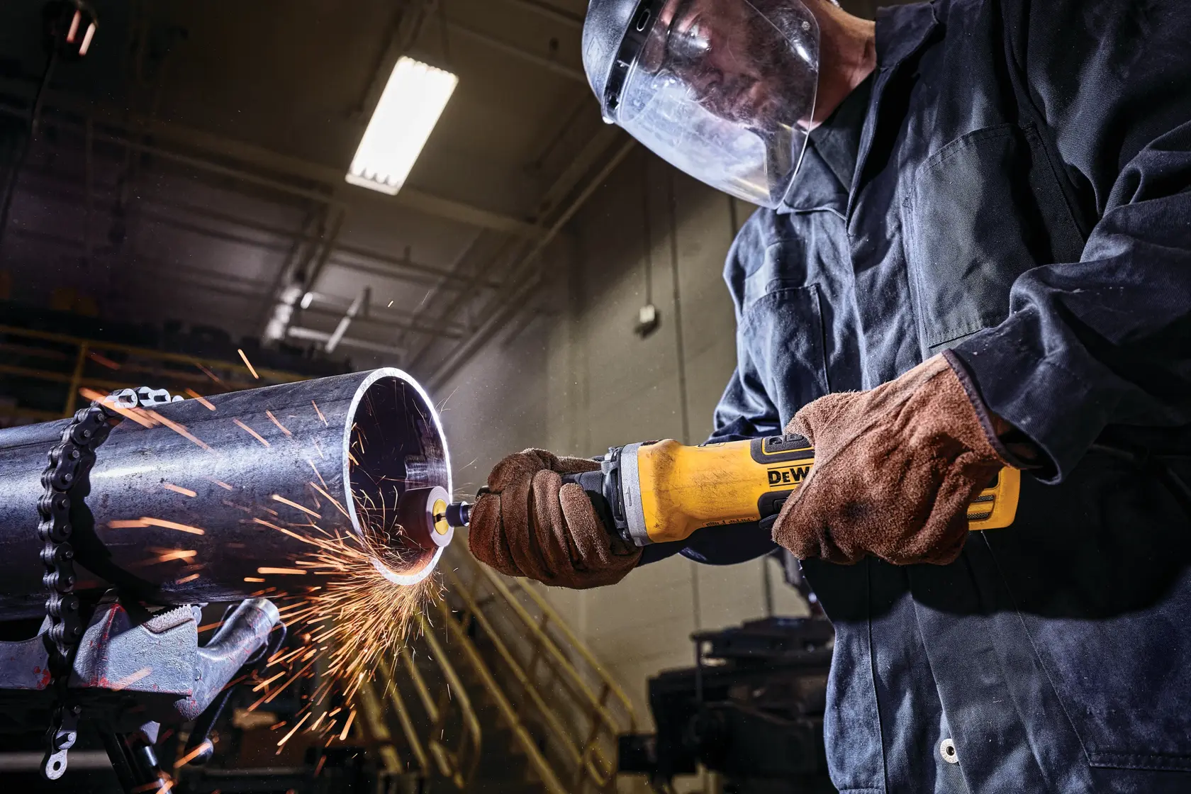 Una persona que lleva guantes protectores y una pantalla facial utiliza una herramienta eléctrica amarilla DEWALT para lijar una tubería de metal, con chispas volando en un taller industrial.