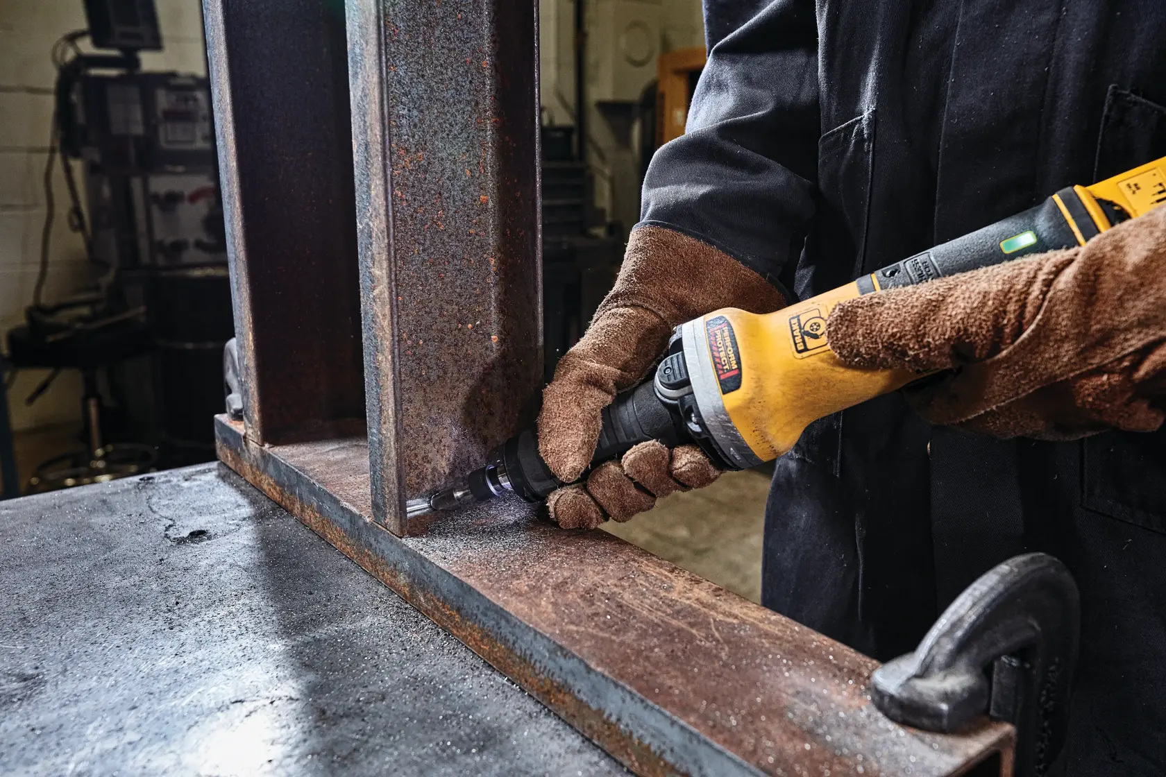 Una persona con guantes marrones utiliza una herramienta eléctrica amarilla y negra DEWALT para pulir o lijar la esquina interior de una viga metálica oxidada en un taller industrial.