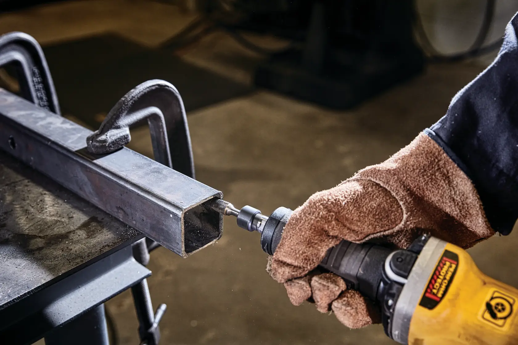 Una persona con guantes de trabajo marrones está usando una herramienta eléctrica amarilla DEWALT sobre un tubo rectangular de metal sujeto a una mesa de trabajo.
