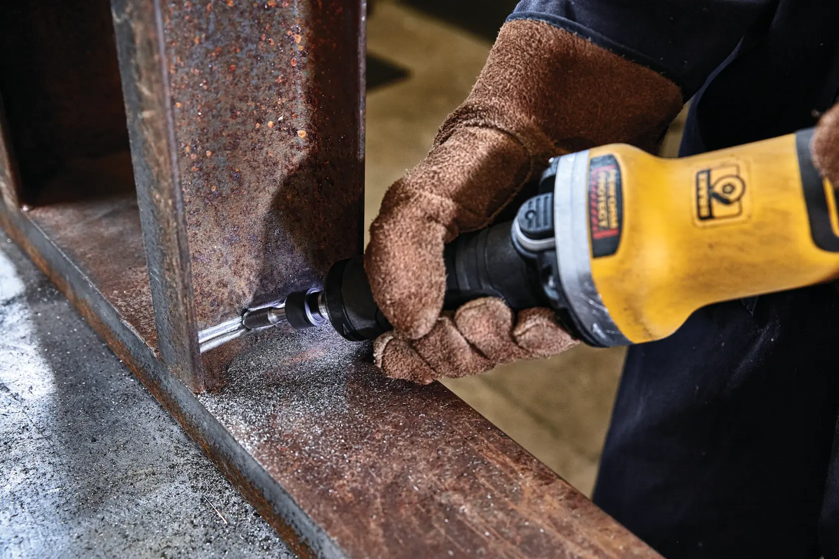 Una persona que lleva guantes de trabajo marrones usa una herramienta eléctrica amarilla DEWALT para esmerilar o pulir la esquina interna de una viga metálica oxidada.