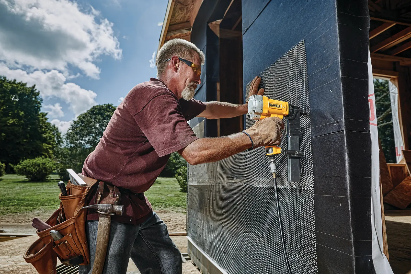 A person using a yellow DEWALT power tool to attach mesh material to the exterior wall of a building under construction. The person is wearing a tool belt and working outdoors on a sunny day.